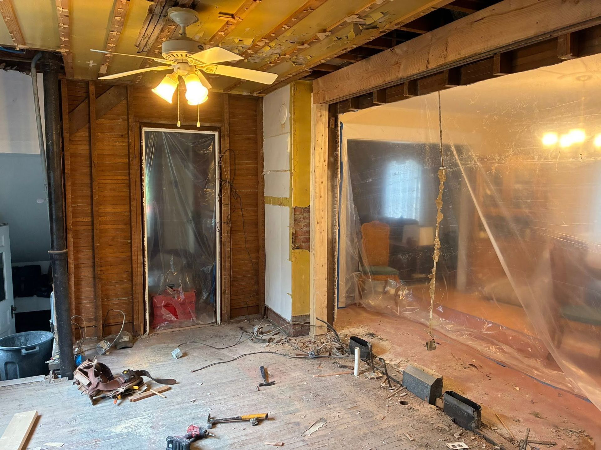 Partially demolished room with exposed studs, plastic sheeting, debris, and a ceiling fan lit overhead
