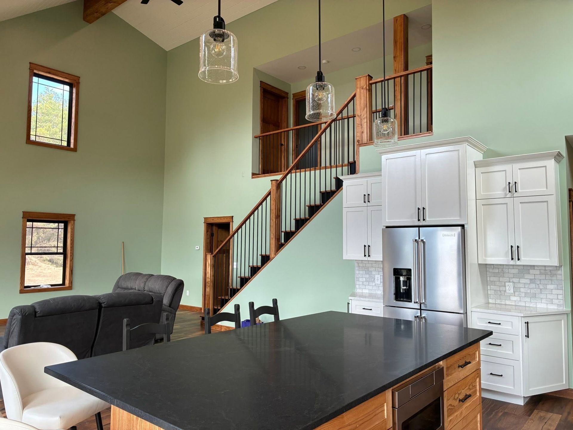 Modern kitchen and living room with dark island, white cabinets, stainless fridge, and staircase.