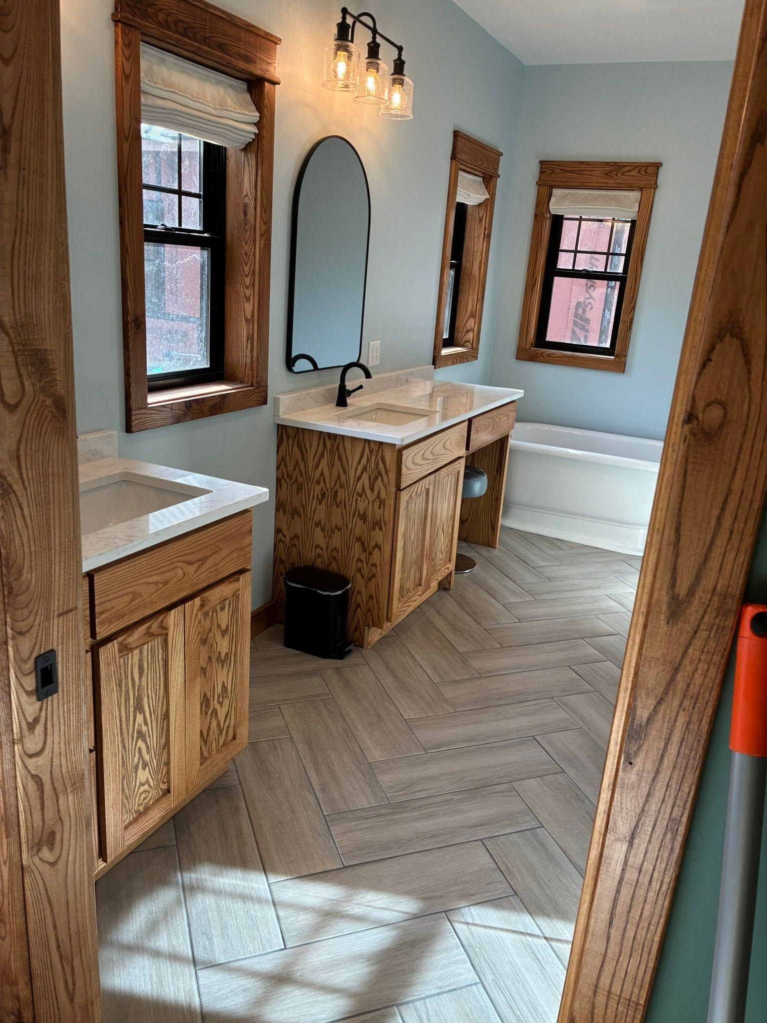 Modern bathroom with wood vanity, large mirror, tiled floor, windows, and a freestanding tub