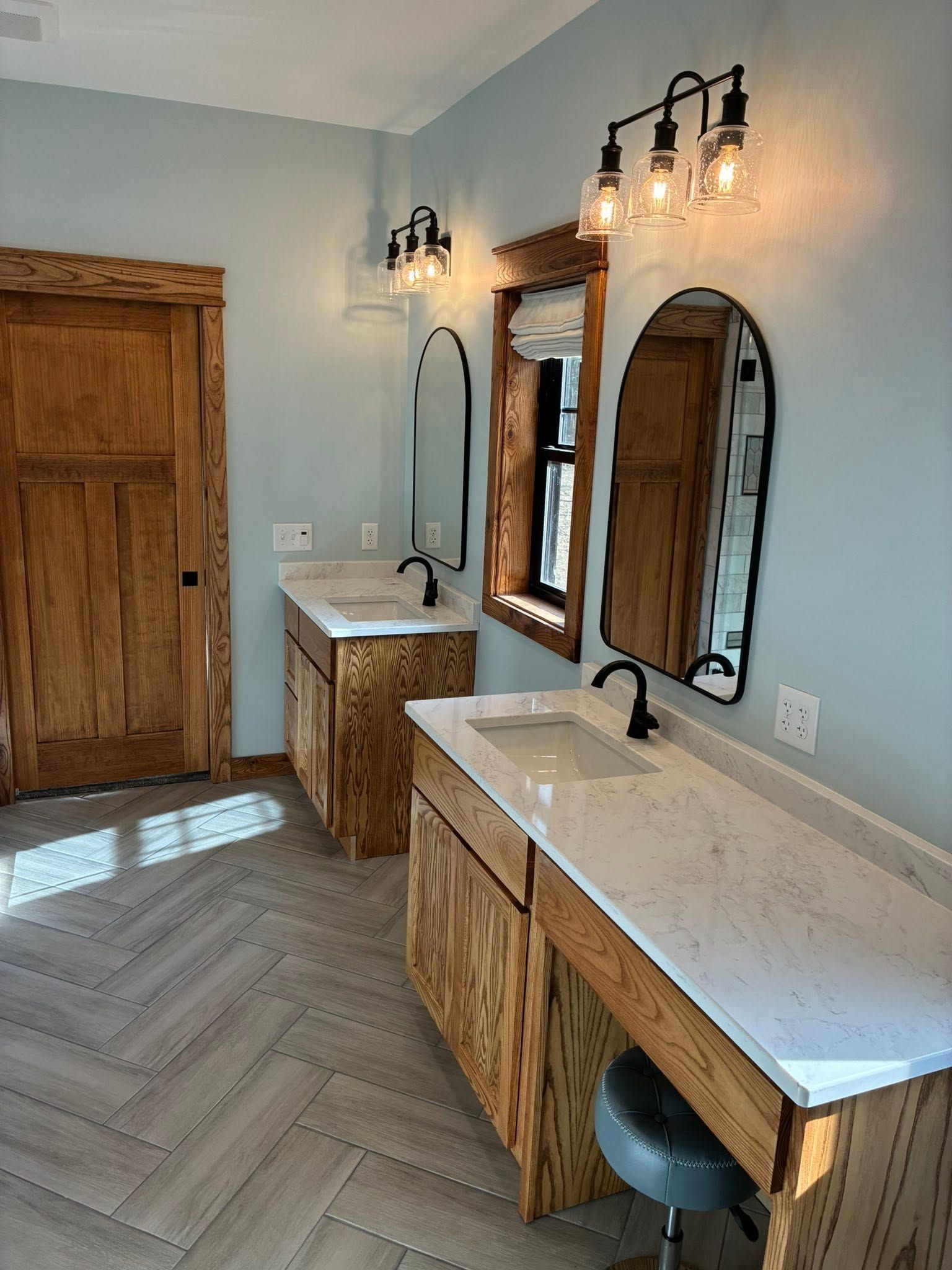 Modern bathroom vanity with wood cabinets, white countertops, arched mirrors, and black sconces.
