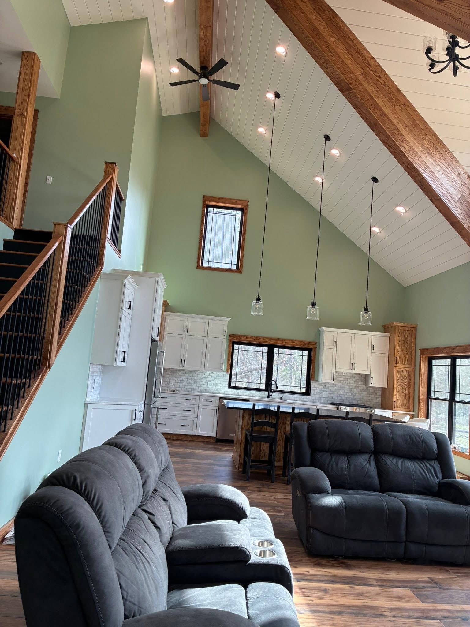 Bright living room with vaulted ceiling, black recliners, staircase, kitchen, and hanging lights