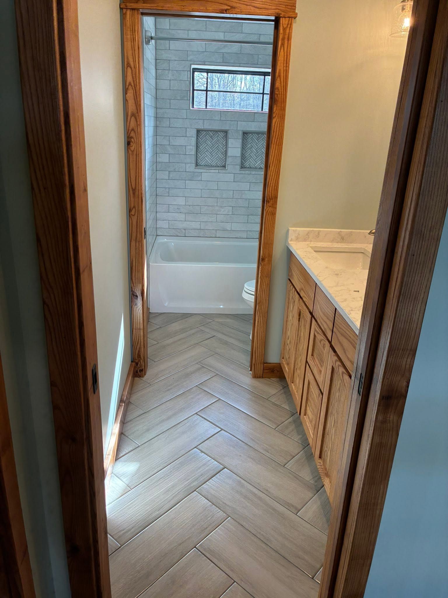 Narrow bathroom hallway with herringbone wood floor, leading to a tub and tiled shower.