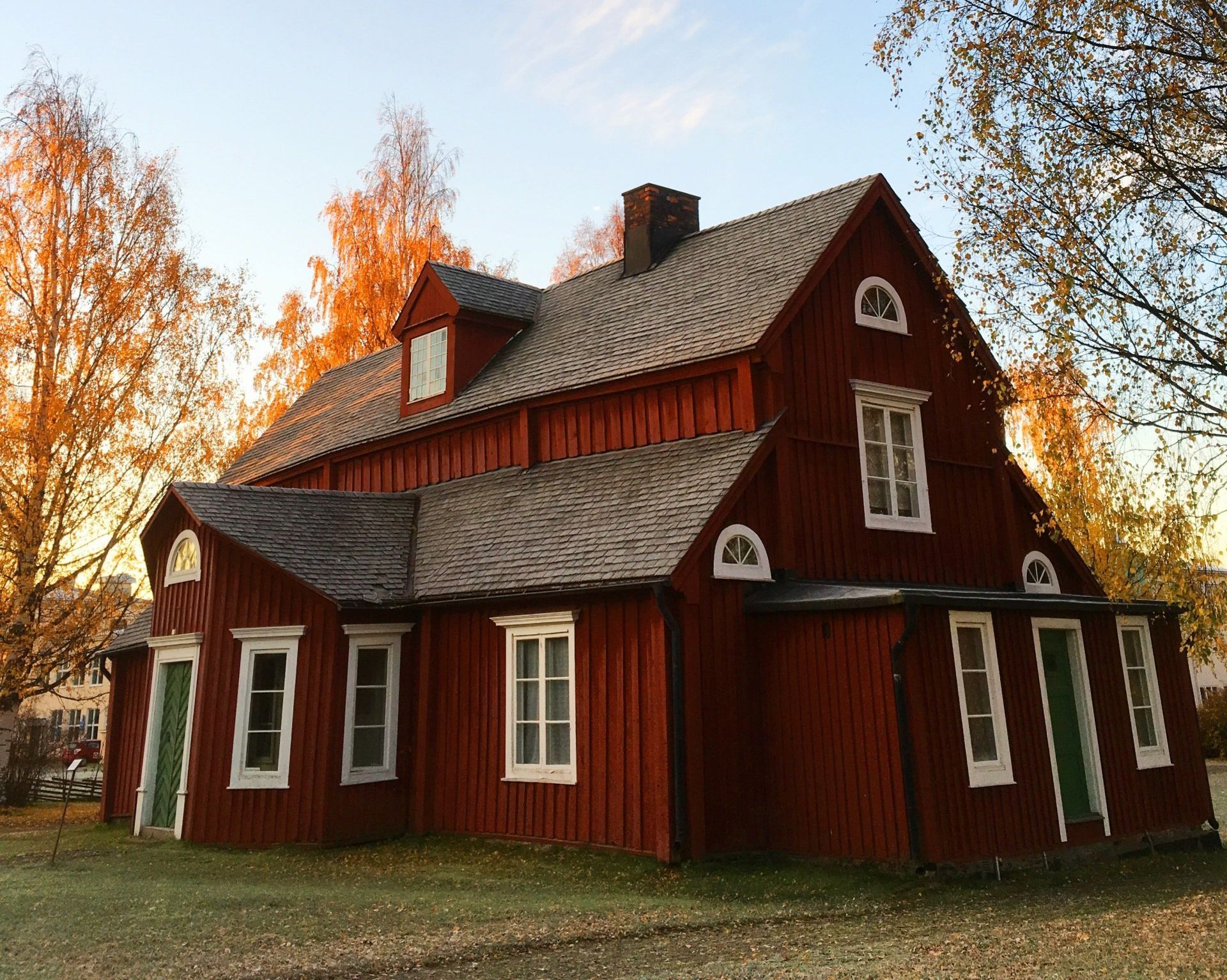 Red wooden house with white-trimmed windows and a dark roof, surrounded by trees with yellow leaves under a blue sky.