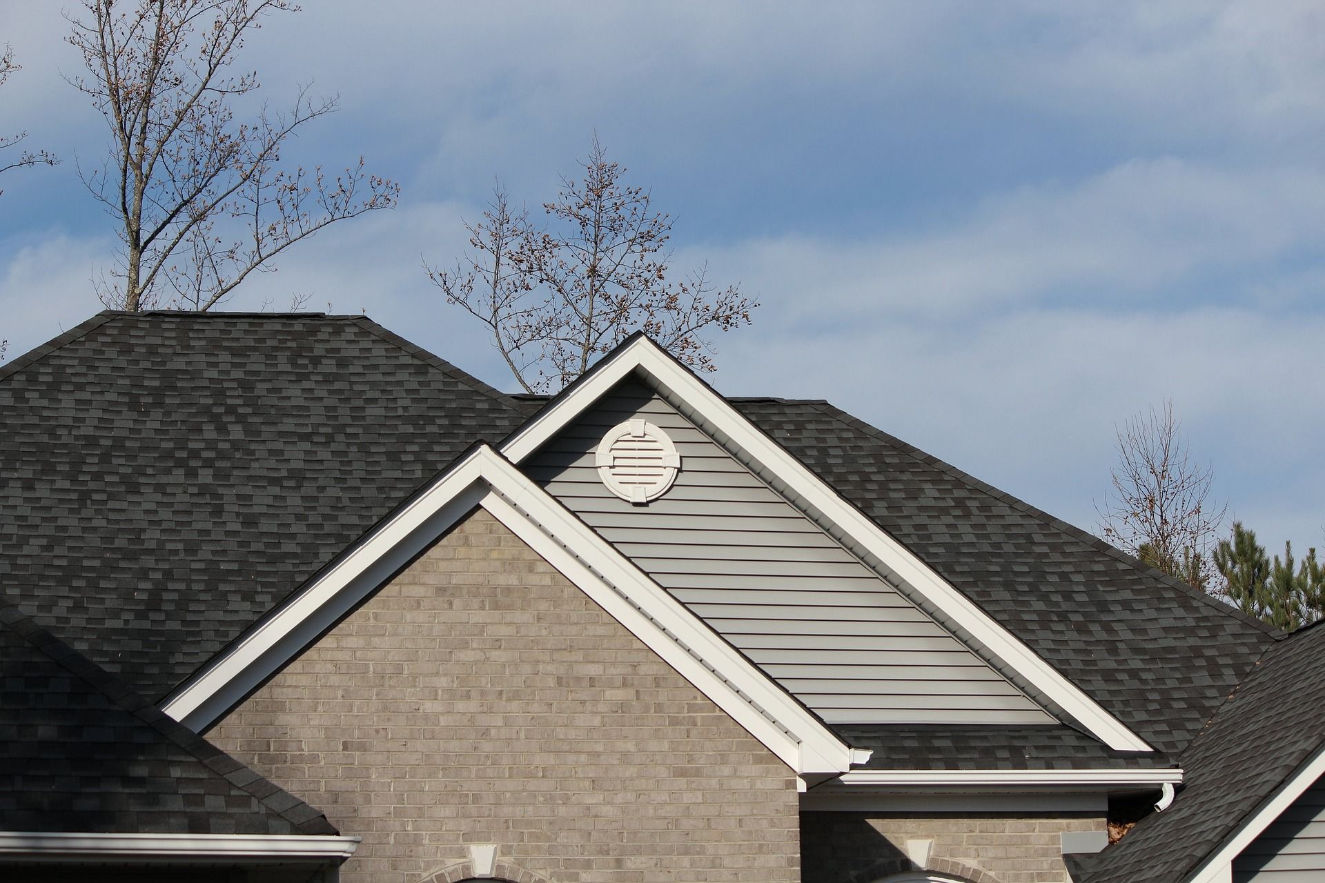 Gray shingled roof with a white-trimmed gable against a blue sky, featuring a small round vent and bare tree branches.