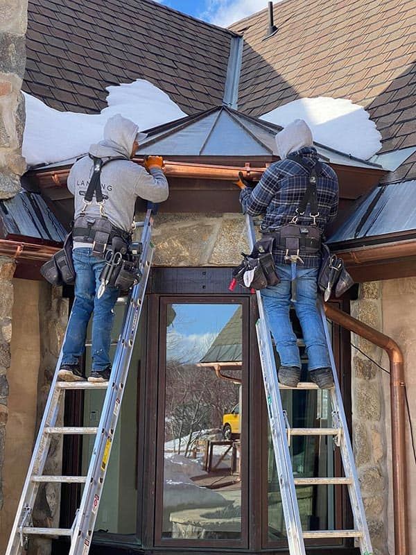 Two workers on ladders repair a damaged roof over a building entrance, with snow on the shingles.