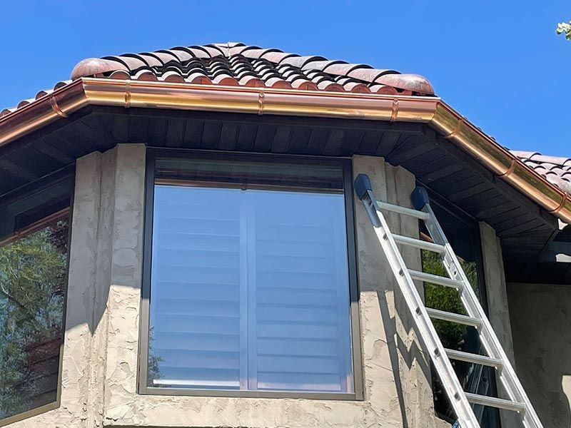 Ladder leaning against a house roof beside a large window under blue sky