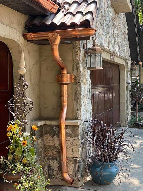 Copper downspout on a stone house beside a lantern, blue planter, and flowers.