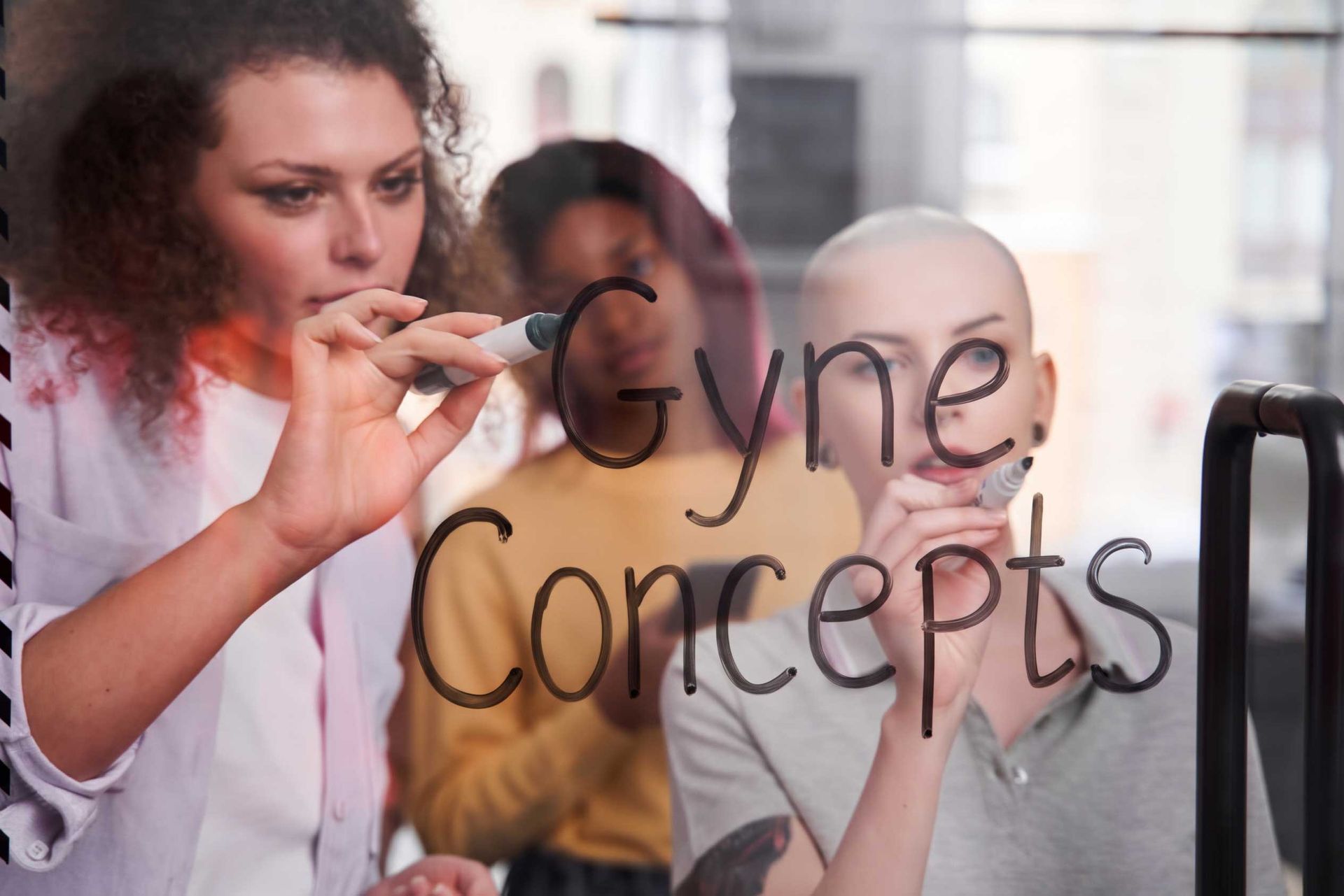 Woman writing “Gyne Concepts” on a glass board in a bright office setting