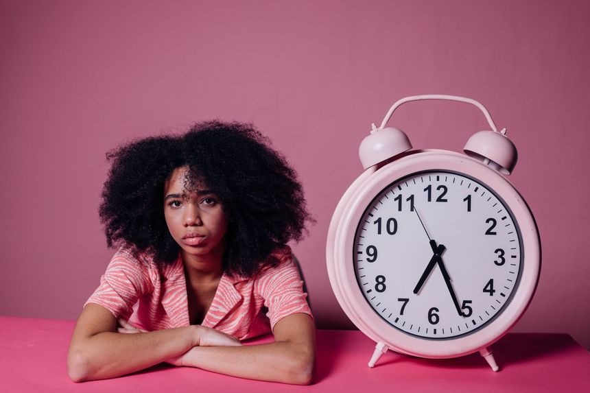Person leaning on a pink surface beside a large pink alarm clock against a pink background