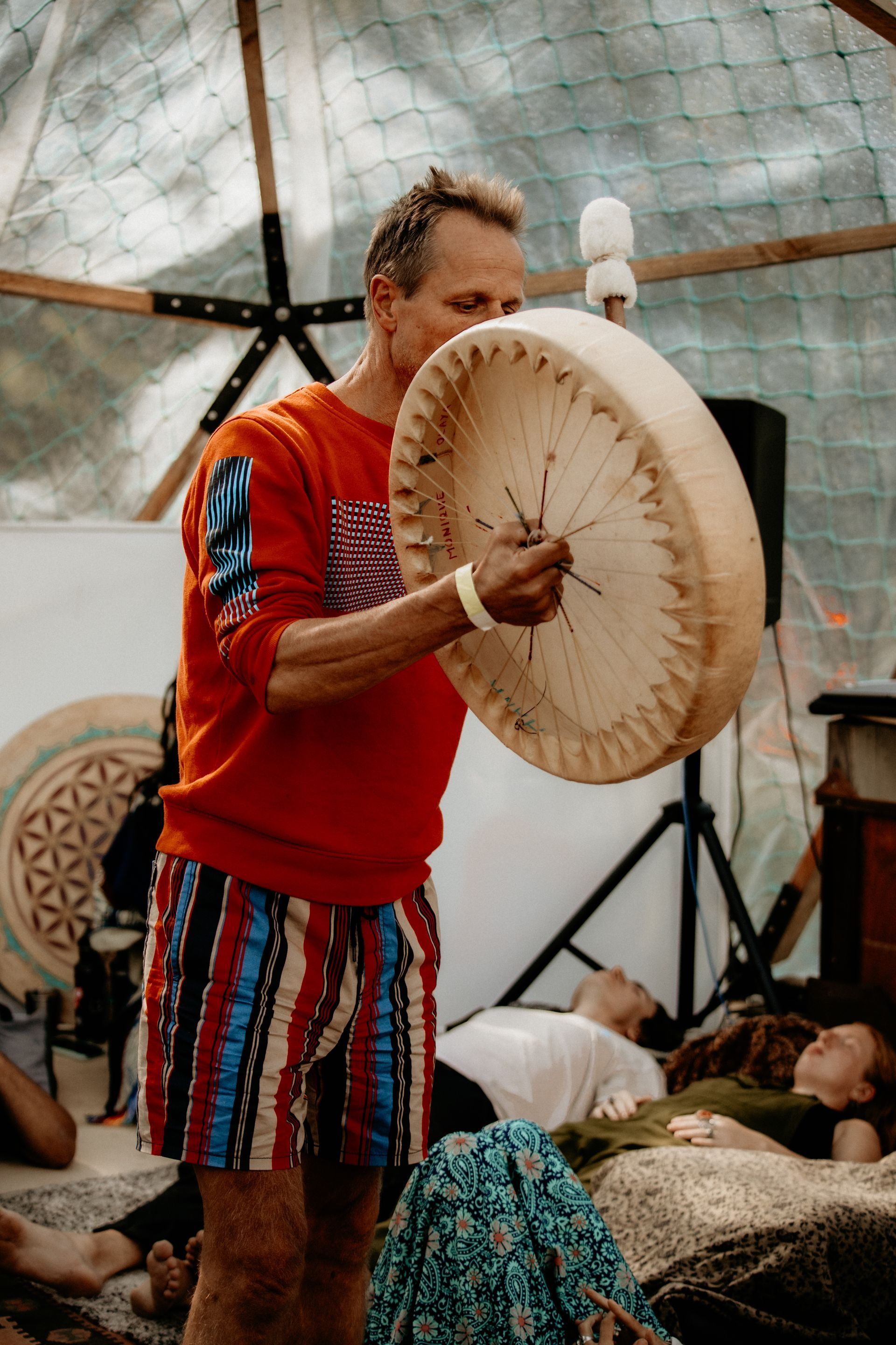 Man in red shirt plays a large drum inside a dome. People are lying down nearby.