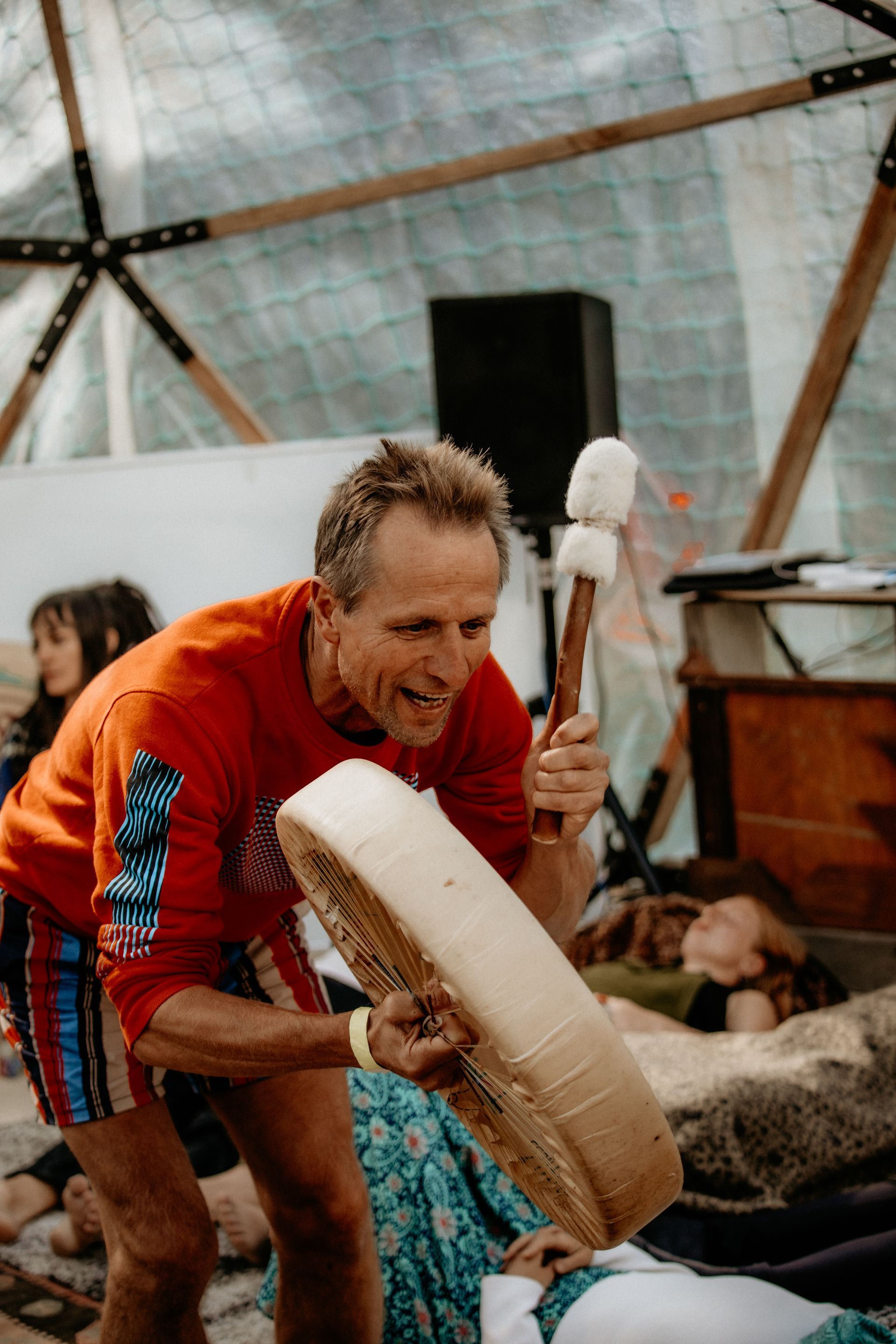 Man in red shirt plays drum, held in front, with drum mallet, inside dome structure.