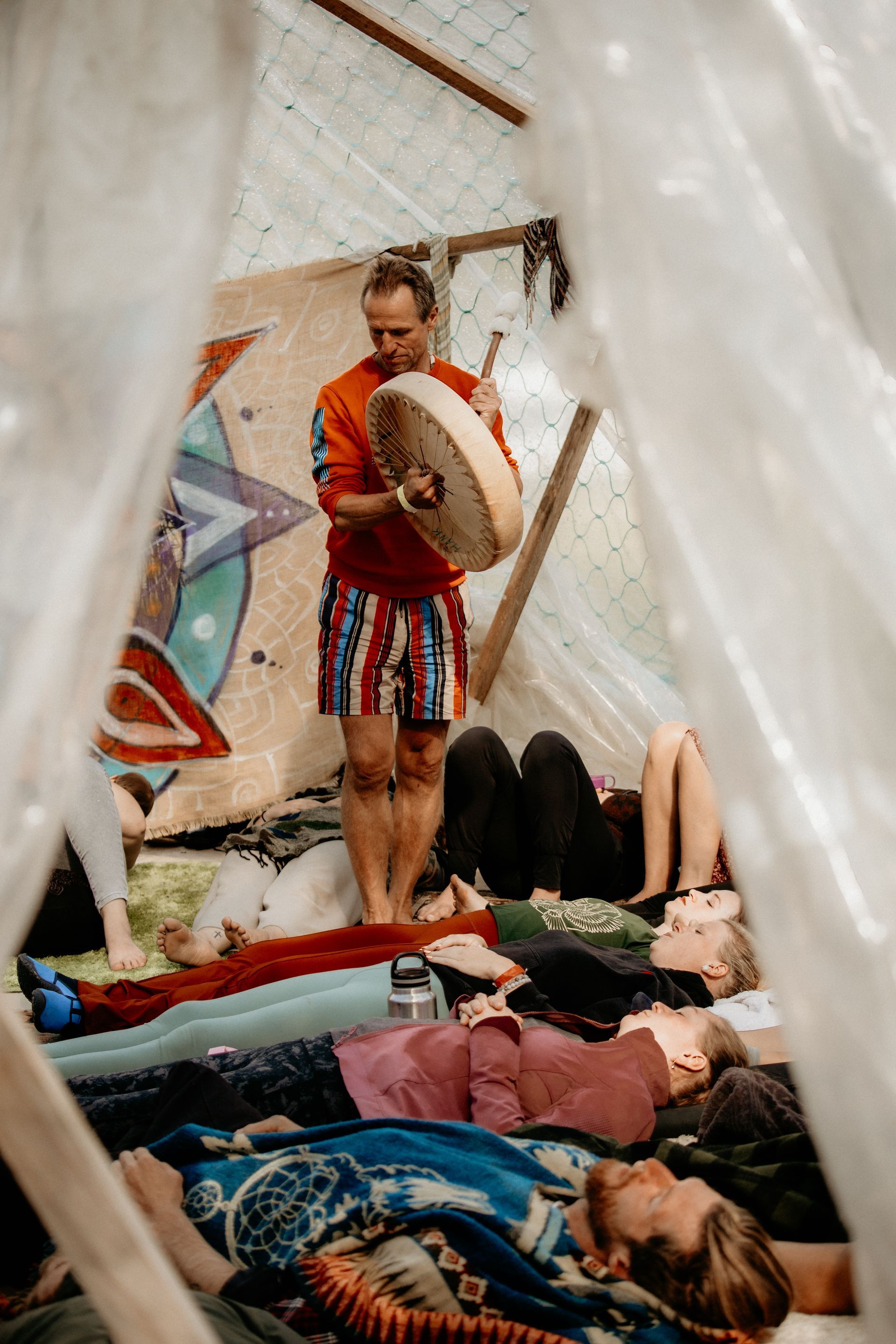 Person drumming over people lying down in a teepee-like structure. Inside, with decorative backdrop and blankets.