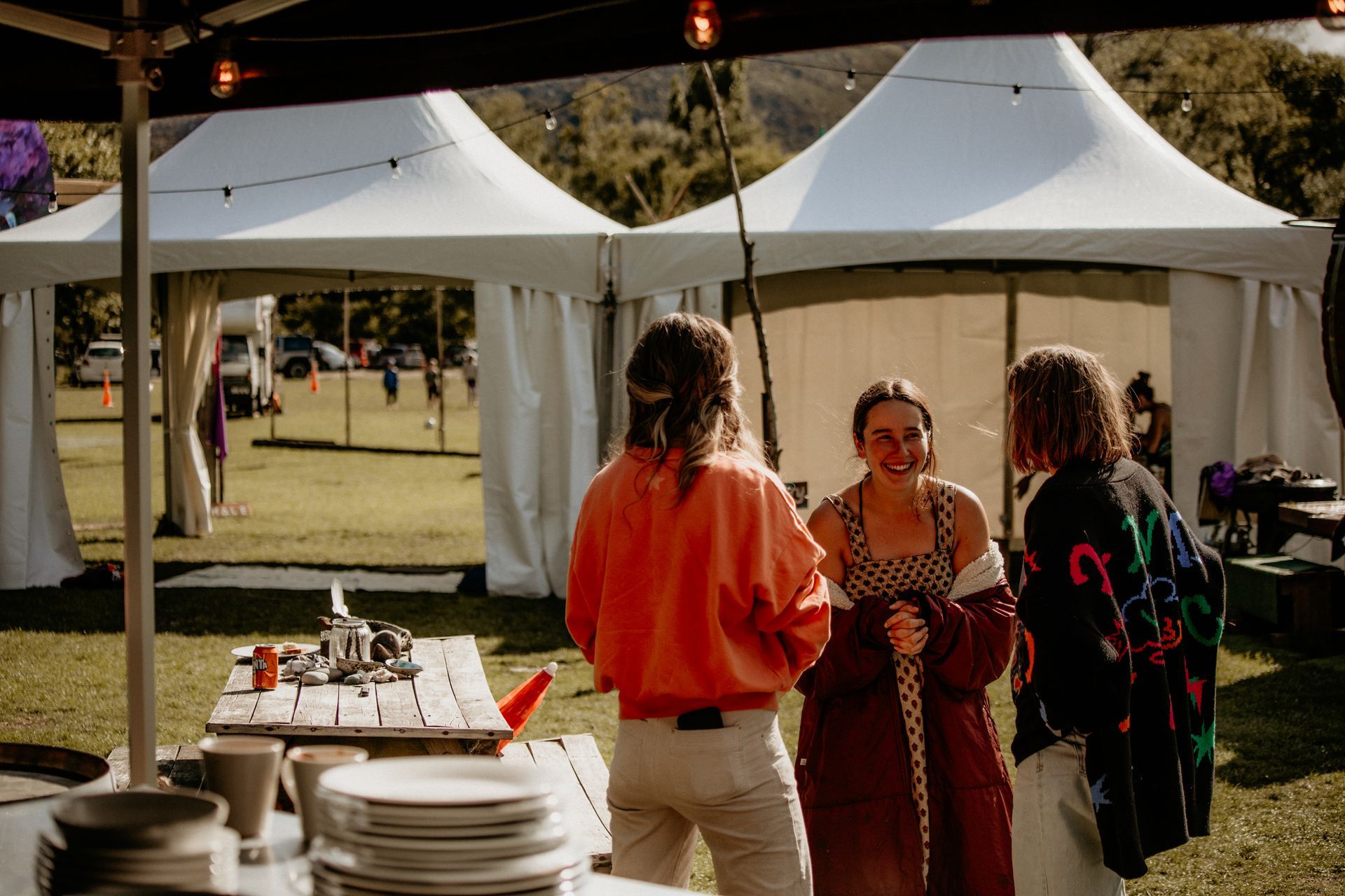 Three people talking by a table with dishes, in a grassy area with tents.