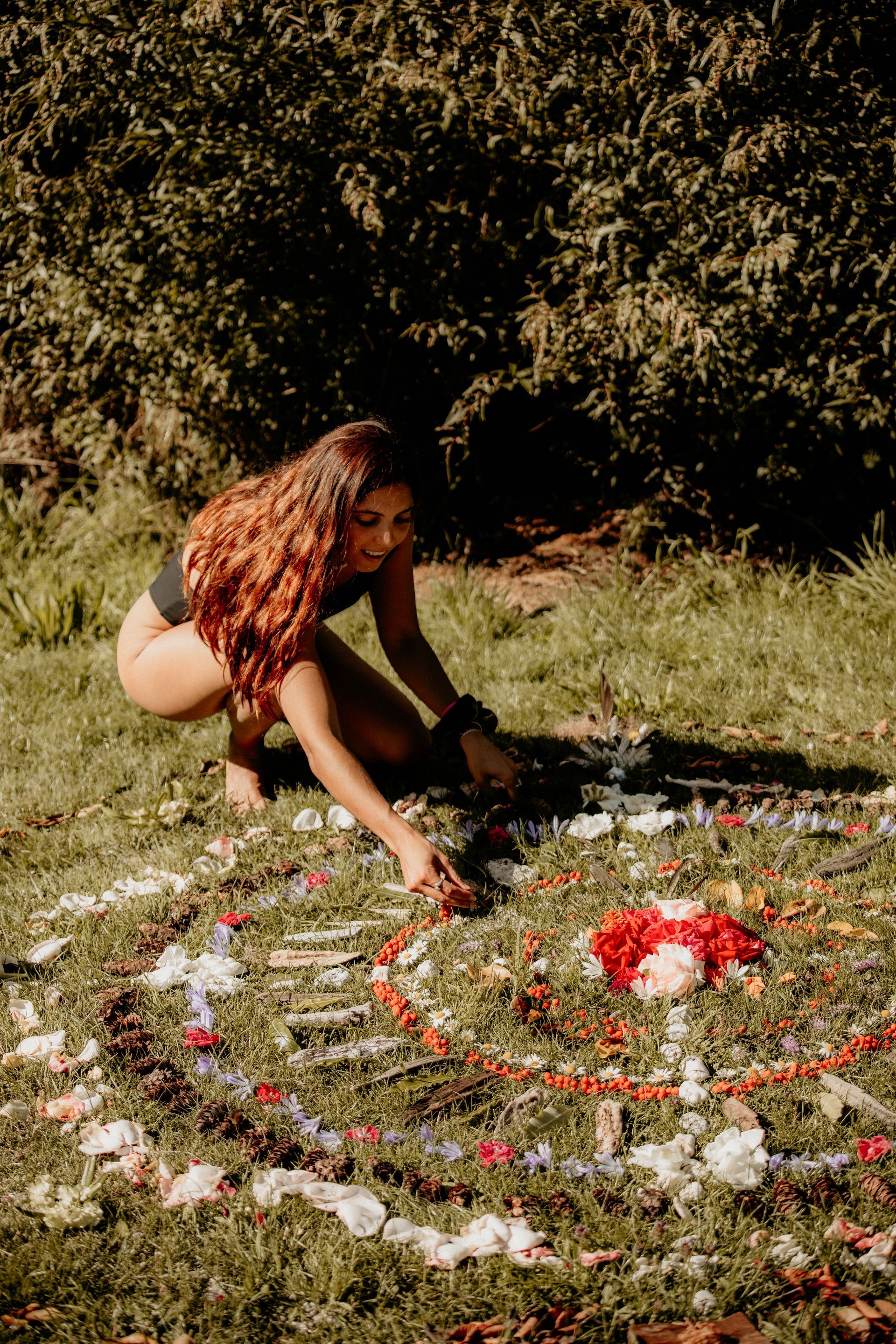 Woman arranging flowers in a spiral on grass. Brown hair, black swimsuit, sunlight, and green foliage background.