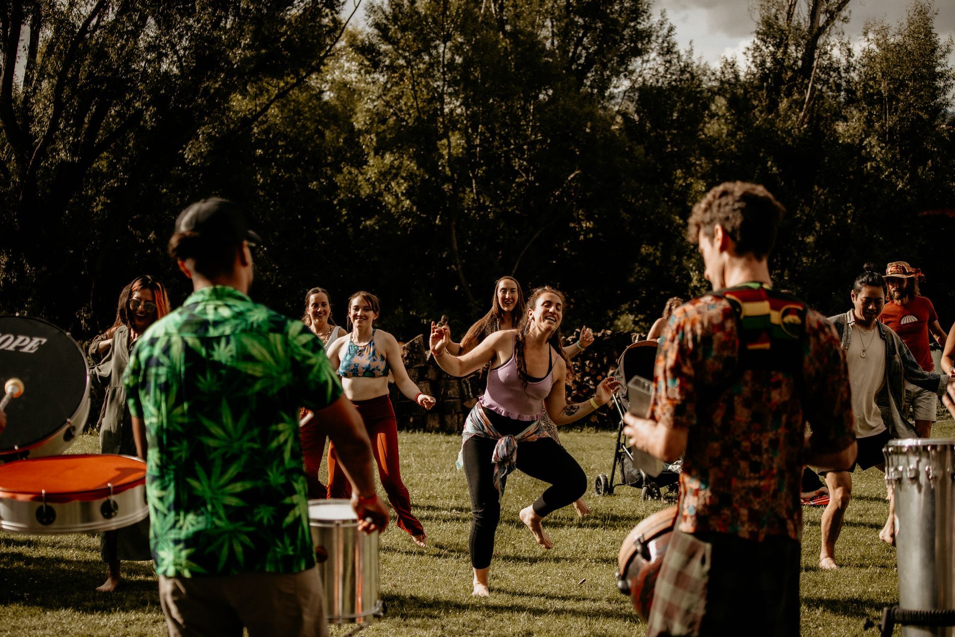 People dancing in a grassy park, a band with drums plays, some wear colorful shirts, happy expressions.