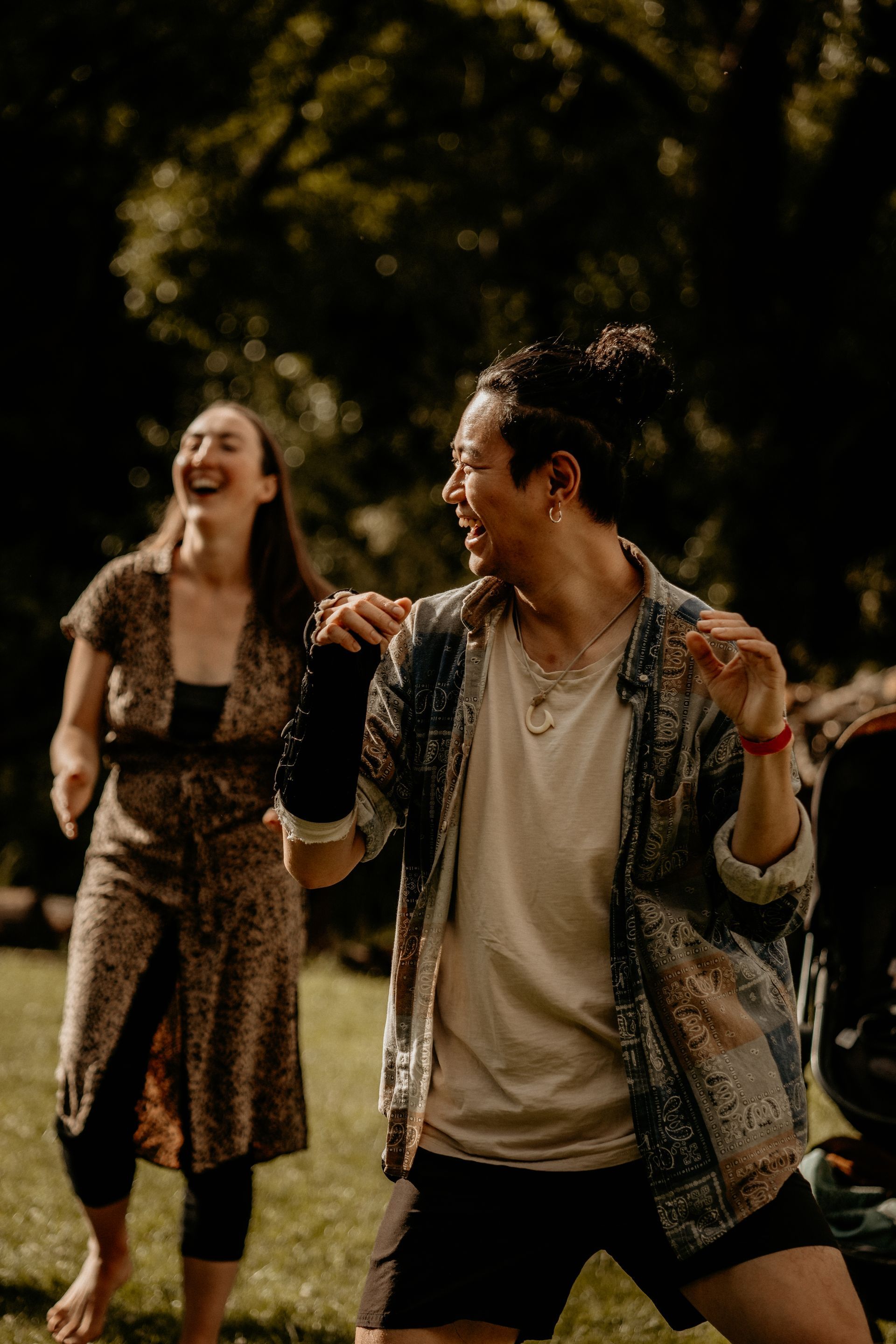Two people laughing and dancing on green grass outdoors. One wears a patterned shirt, the other a long dress.