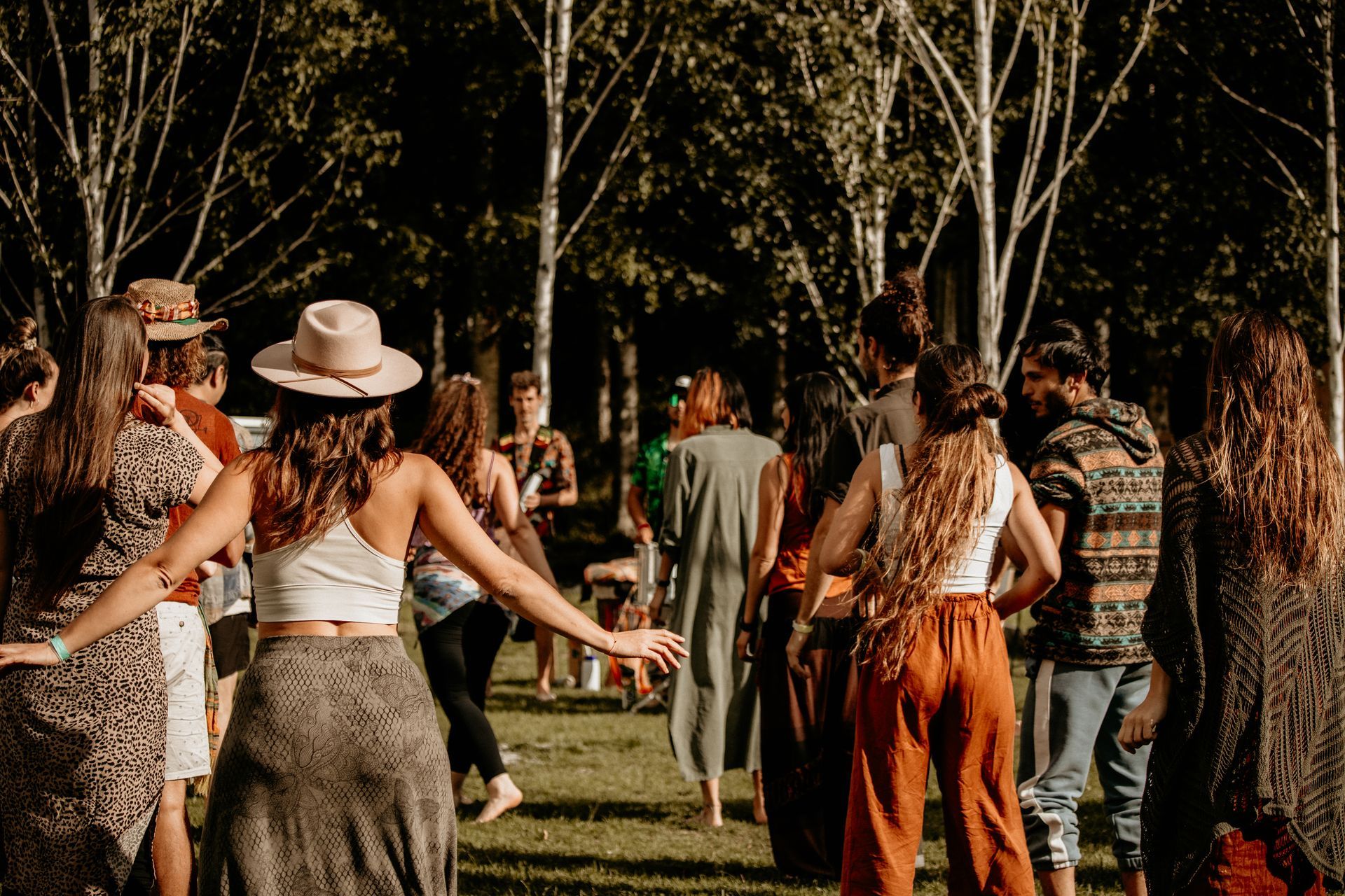 People dancing outdoors in a circle, trees in the background. Sunny day, casual clothing.