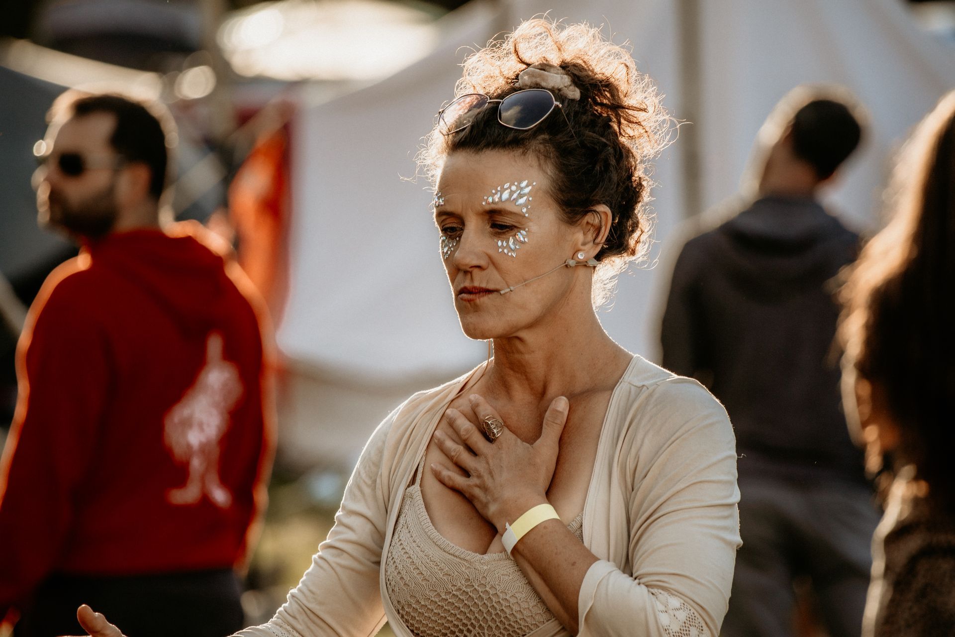 Woman with glitter on her face, hands on chest, eyes closed. Outdoor event, other people blurred in background.