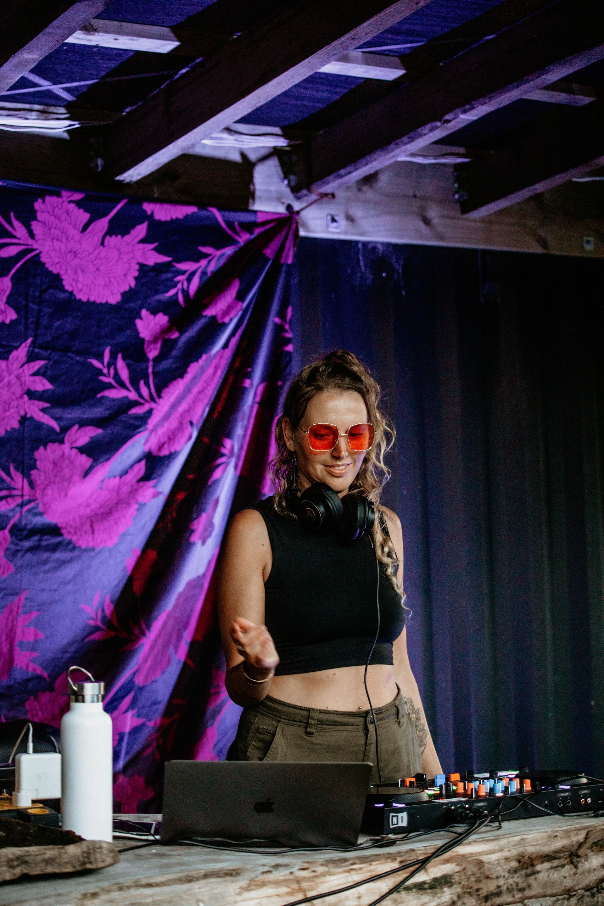 DJ smiling, wearing red glasses and headphones, at a booth with equipment and purple backdrop.
