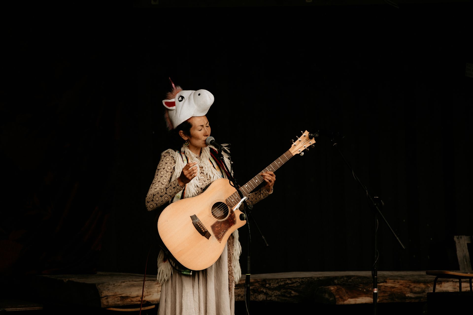 Person on stage singing and playing acoustic guitar, wearing a unicorn head. Black background.