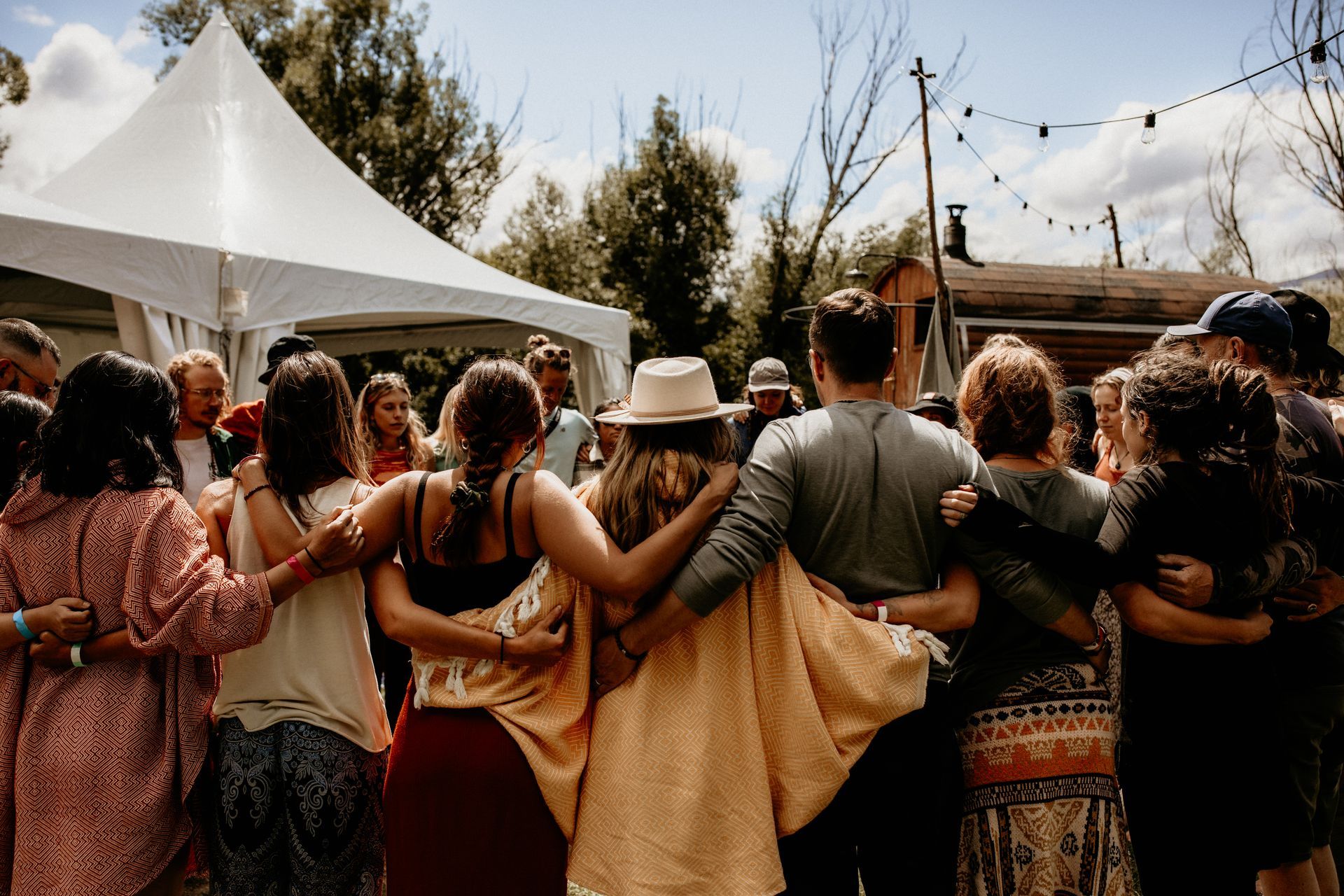 People with arms around each other in a circle, outdoors near a tent and trees.