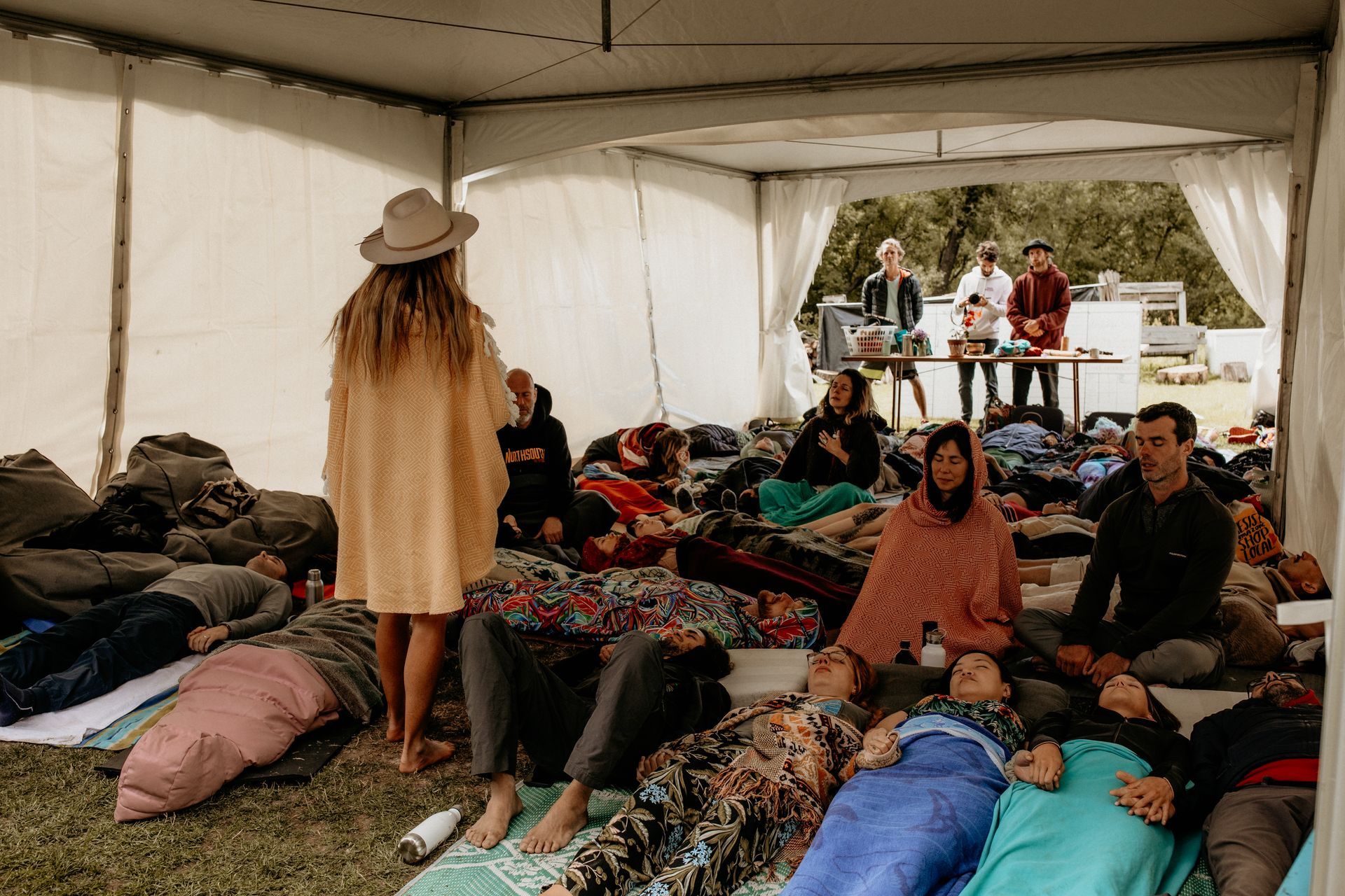 People lying down in a tent on grass, with a person standing, possibly leading a session.