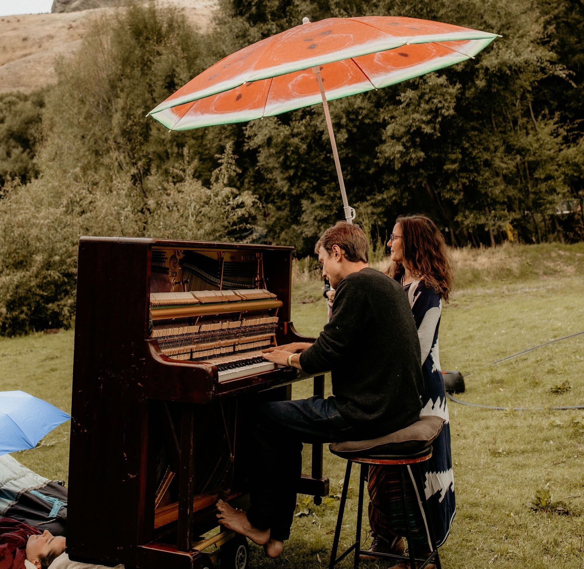 Man plays piano outdoors under an umbrella, a woman stands nearby.