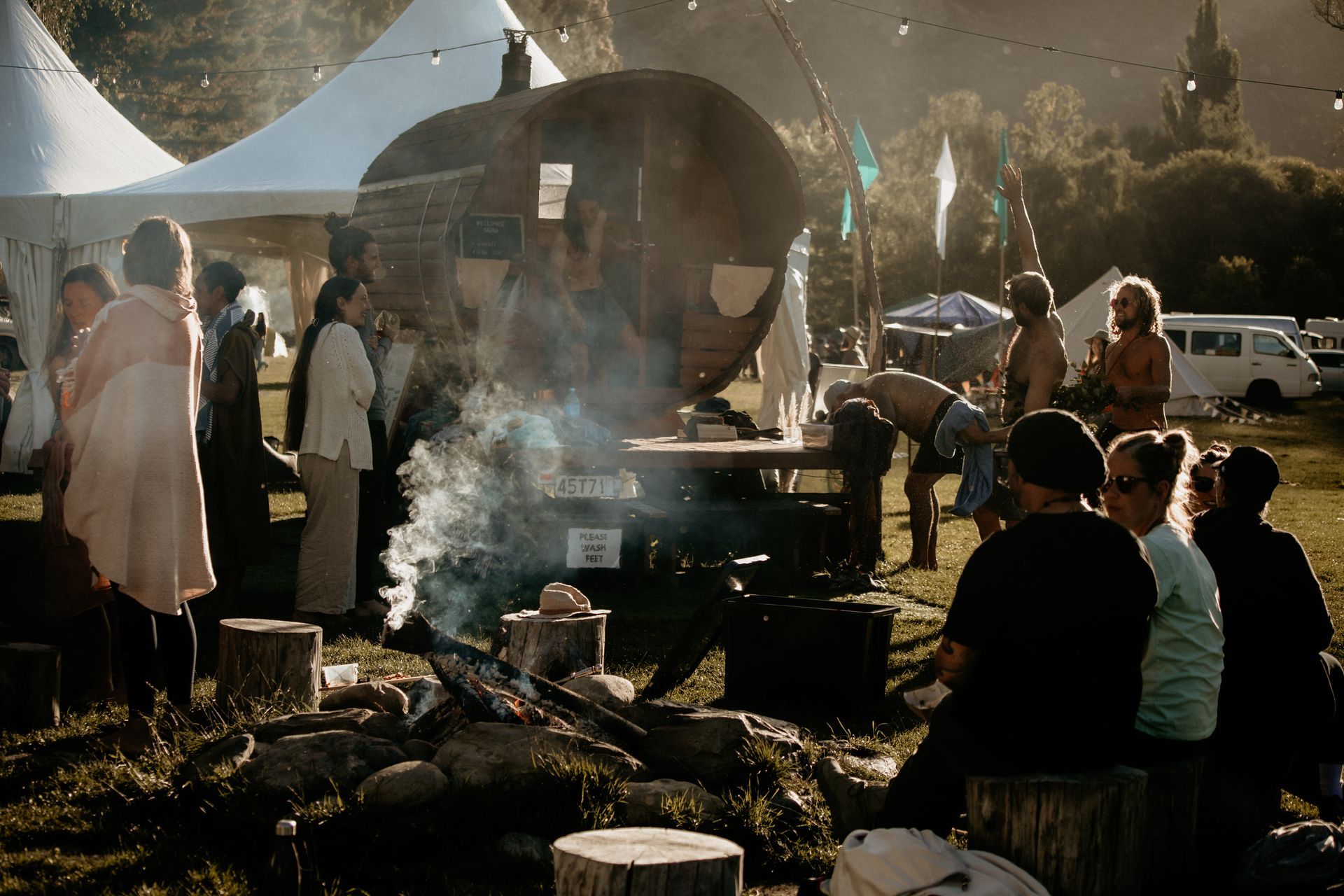 People gather around a fire near a caravan at an outdoor festival. Smoke rises, tents in the background.