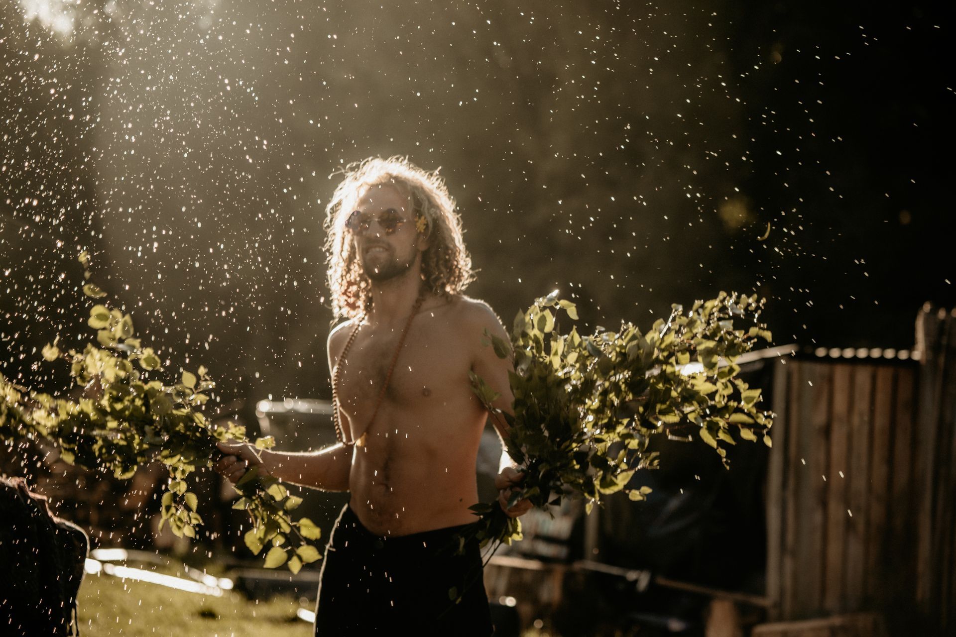 Man with long hair and sunglasses, shirtless, holding leafy branches, standing in sunlight with water droplets.