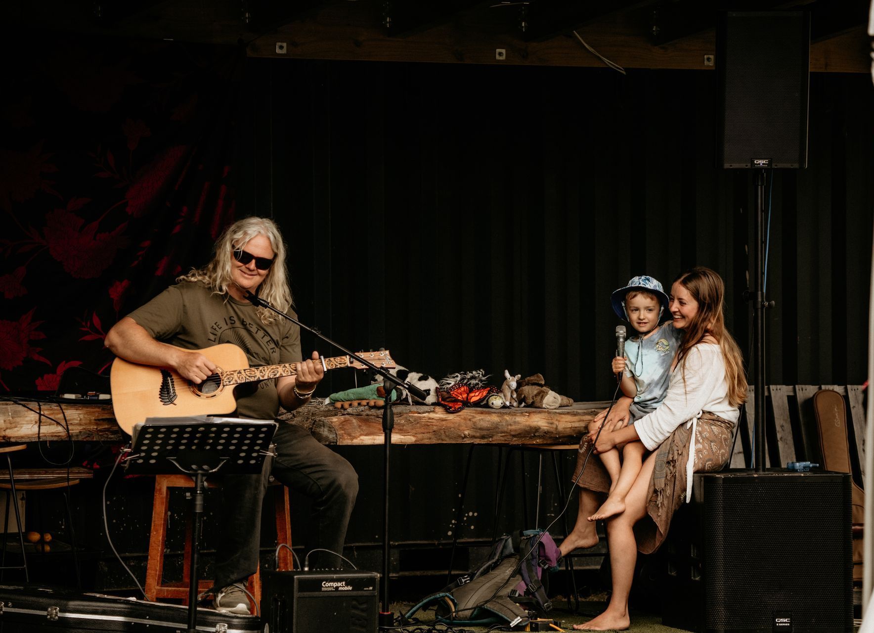Musician playing guitar on stage; a young person and child watch. Black backdrop.