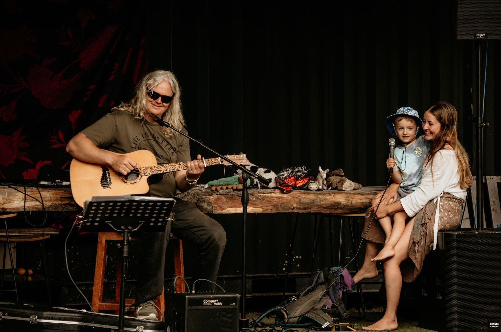 Man playing acoustic guitar on stage, children watching, rustic table.