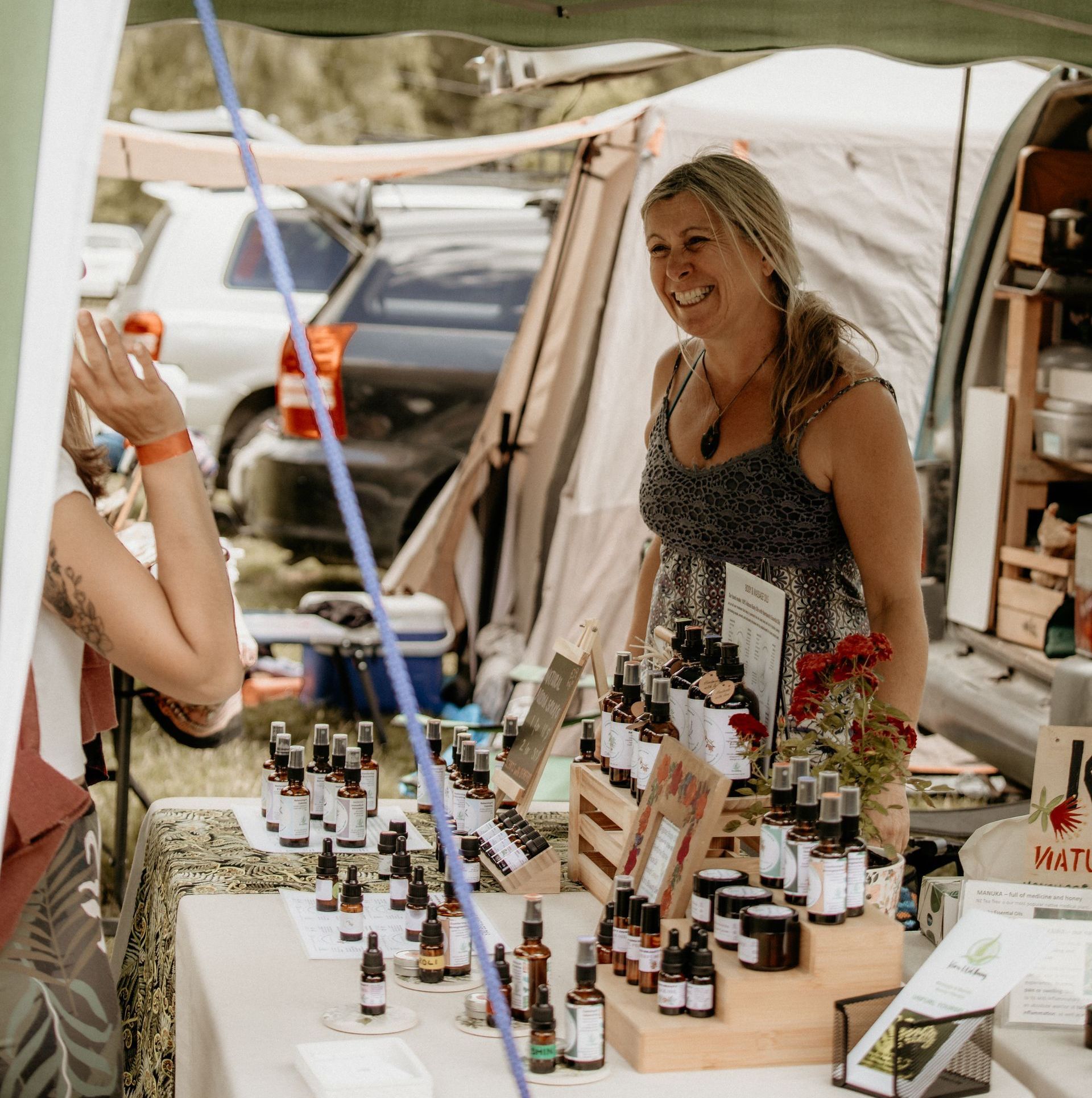 Woman smiling at a booth, selling lotions and sprays at an outdoor market.