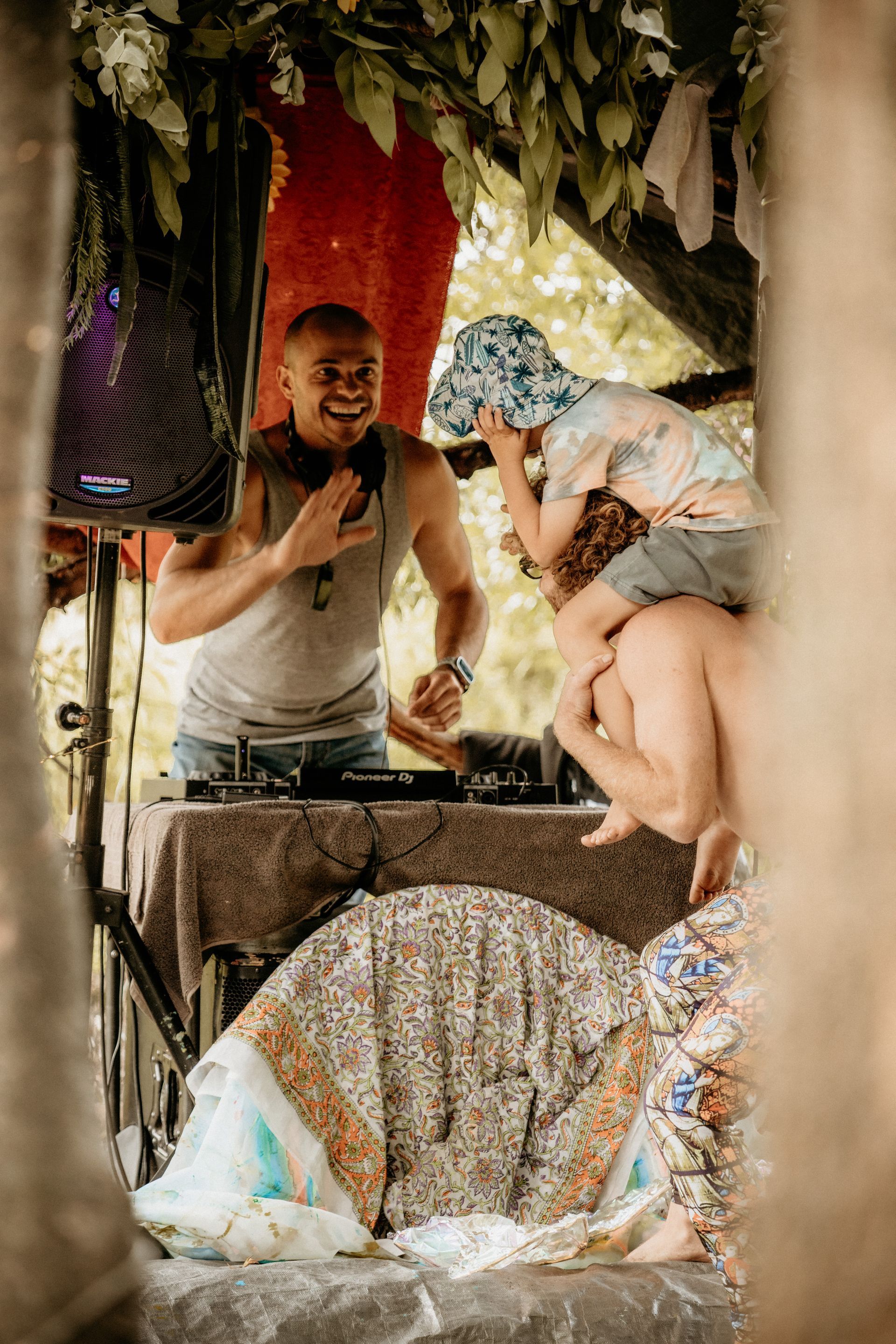 DJ smiles, plays music at outdoor event. Child sits on a person's shoulders, near speaker. Party setting.