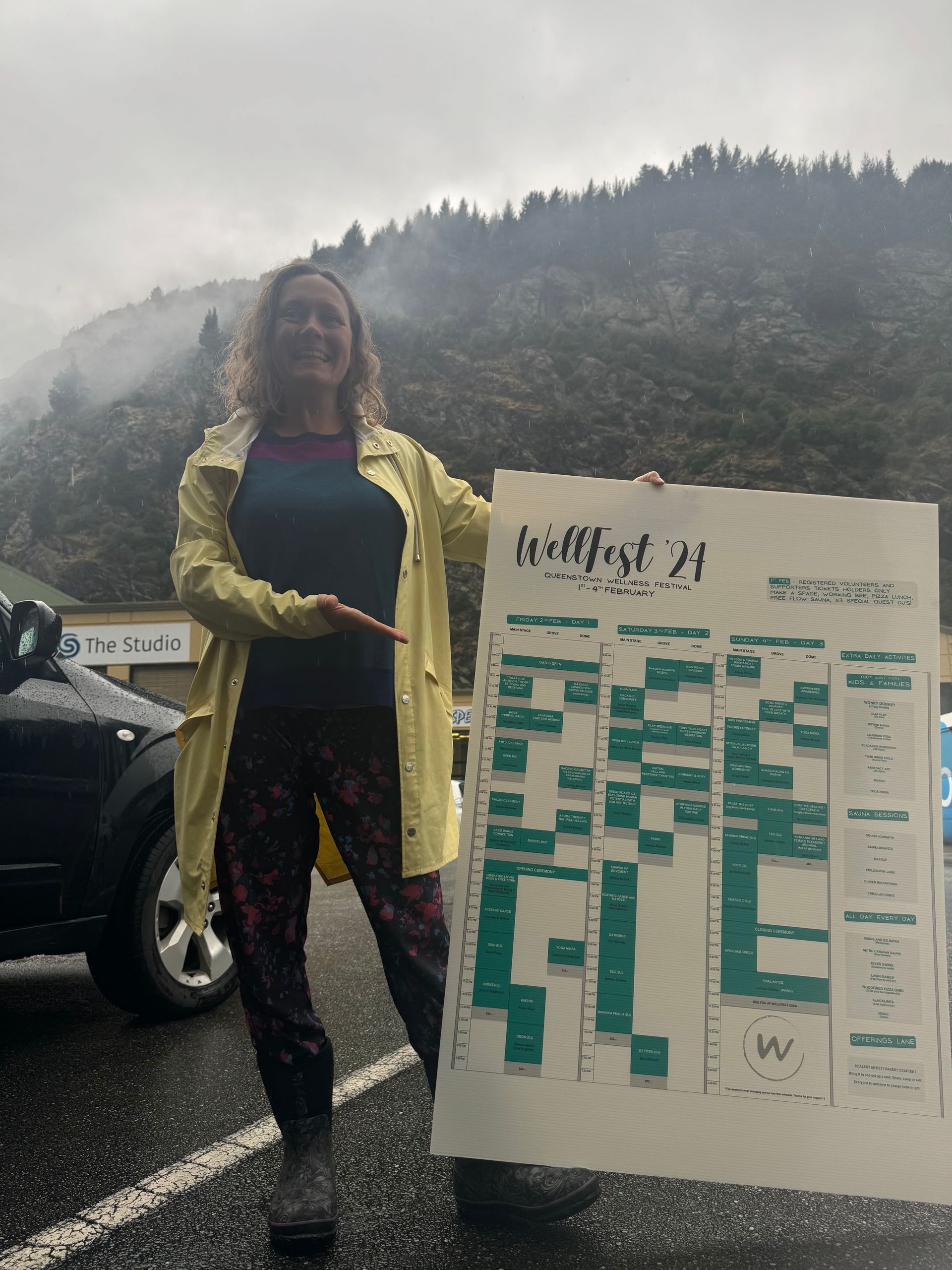 Woman in yellow raincoat holding a schedule poster in front of a mountain.