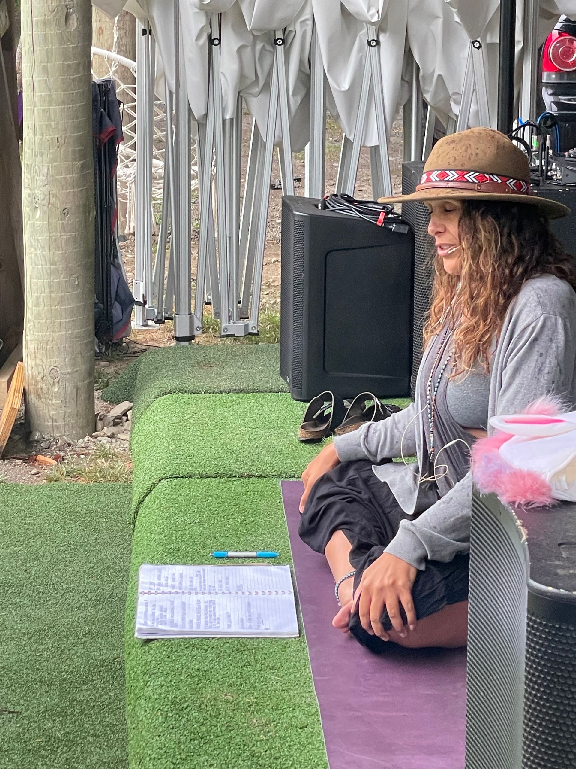 Woman in hat meditates outdoors on a yoga mat, near speakers, clipboard, and green turf.