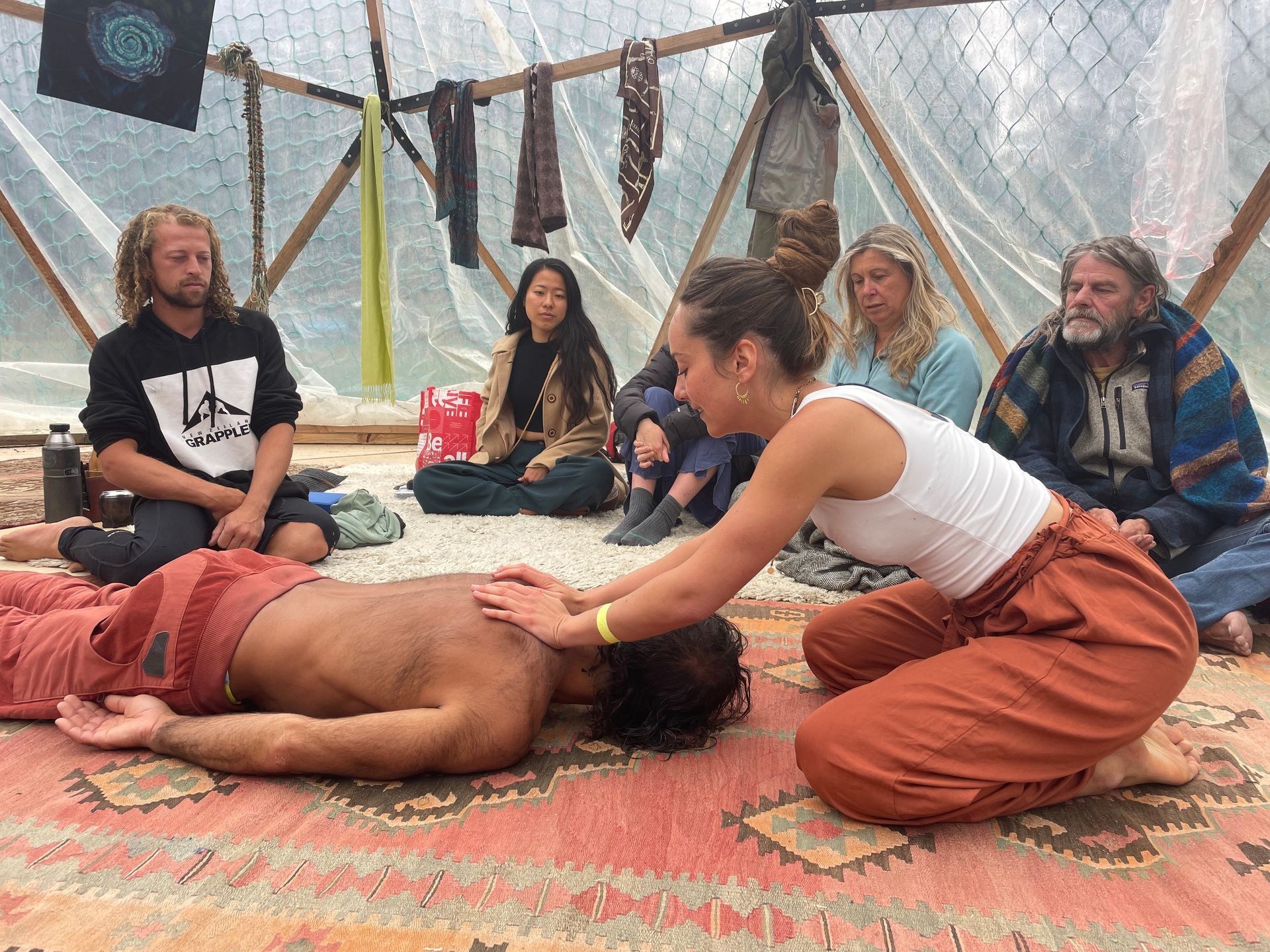 A woman giving a massage to a person lying on a rug. Others observe. Indoors with a translucent dome roof.
