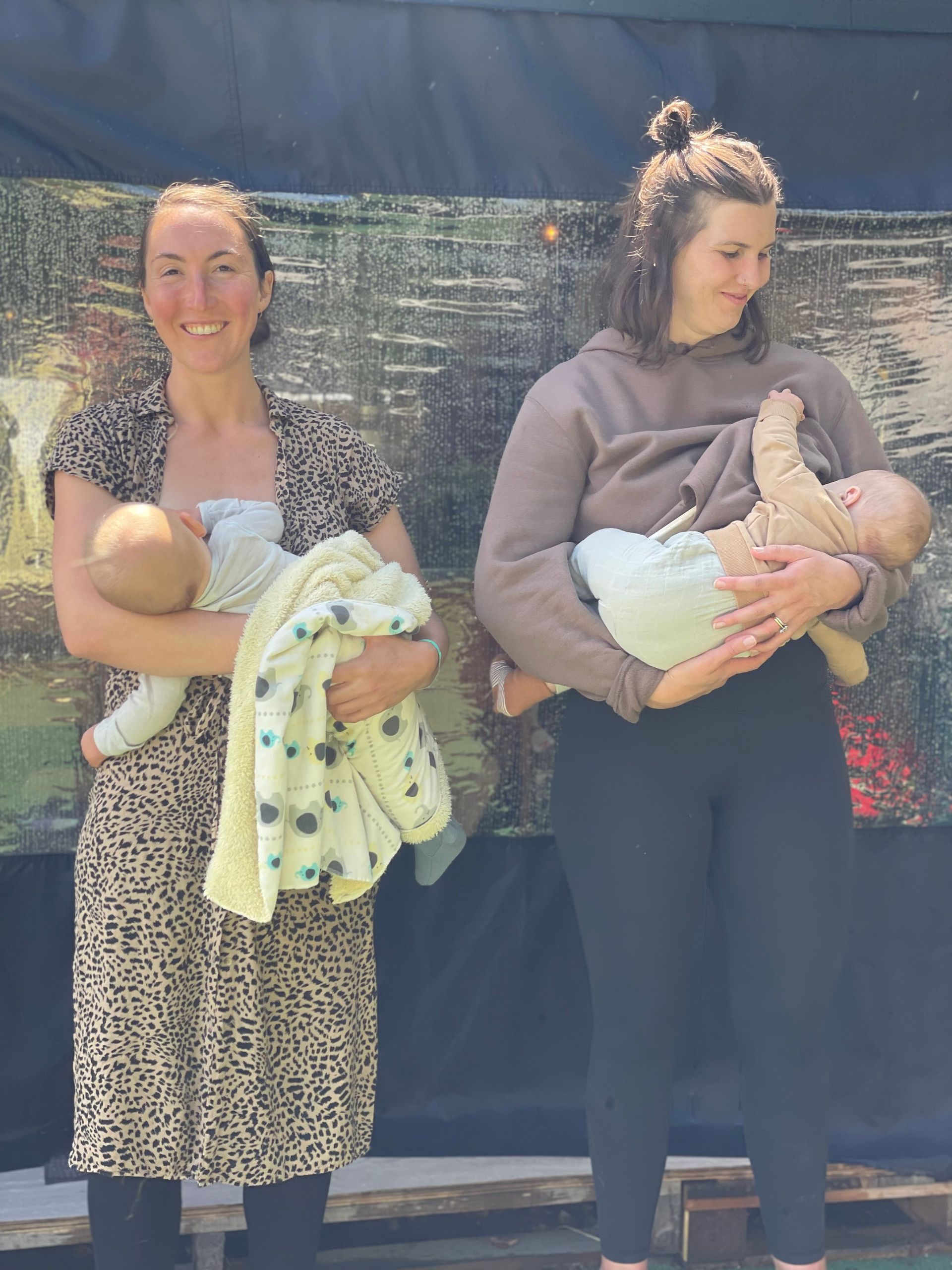 Two women breastfeeding babies outdoors, smiling.