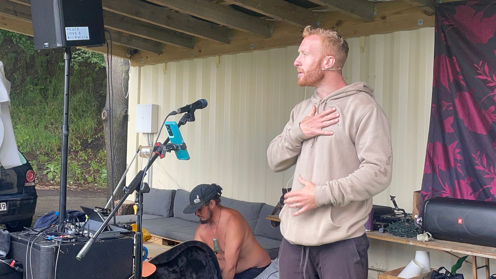Man singing into a microphone, hand on chest, outdoor setting. Another person seated nearby.