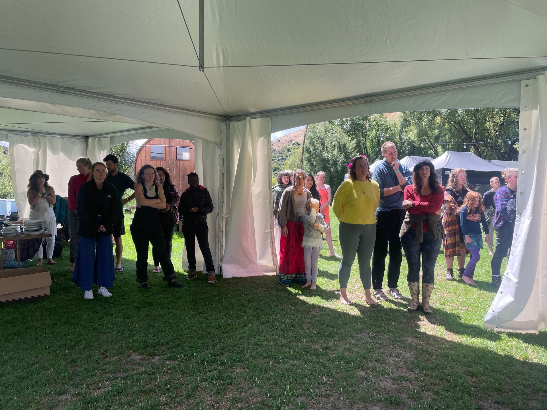 People gather under a white tent on a grassy lawn. Some look towards something outside the tent.