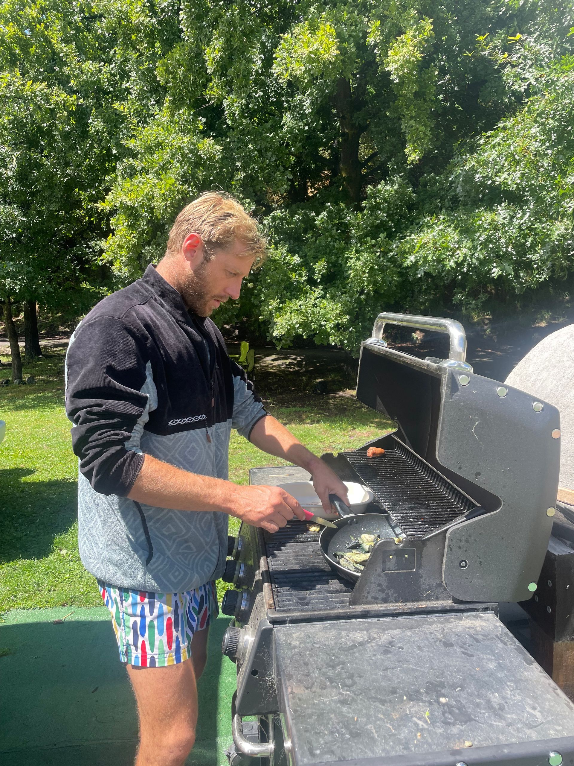 Man grilling outdoors, wearing a jacket and colorful shorts, surrounded by trees.