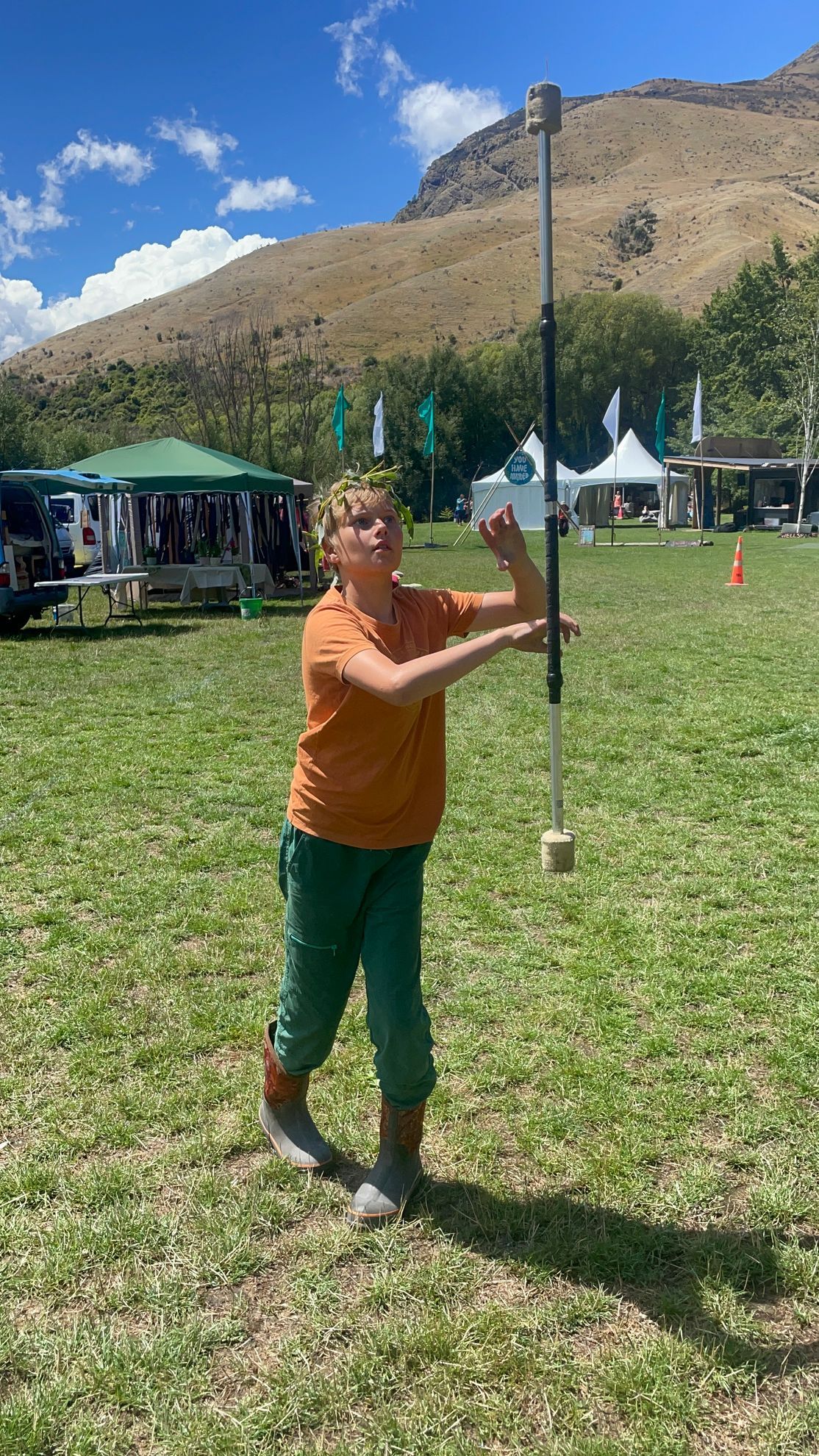 Person in green pants and orange shirt balances a staff outdoors, with a mountain backdrop.