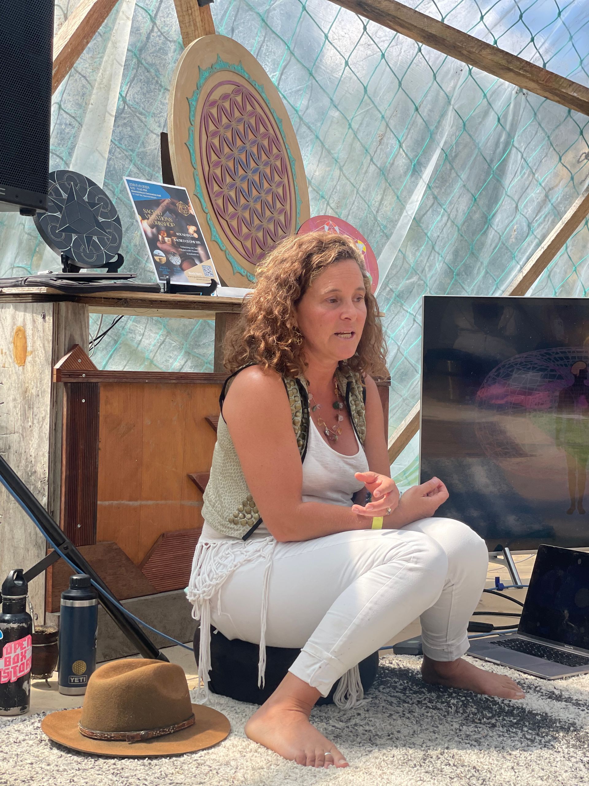 Woman with curly hair sits cross-legged, speaking. She wears white and a vest, in a dome with art and tech.