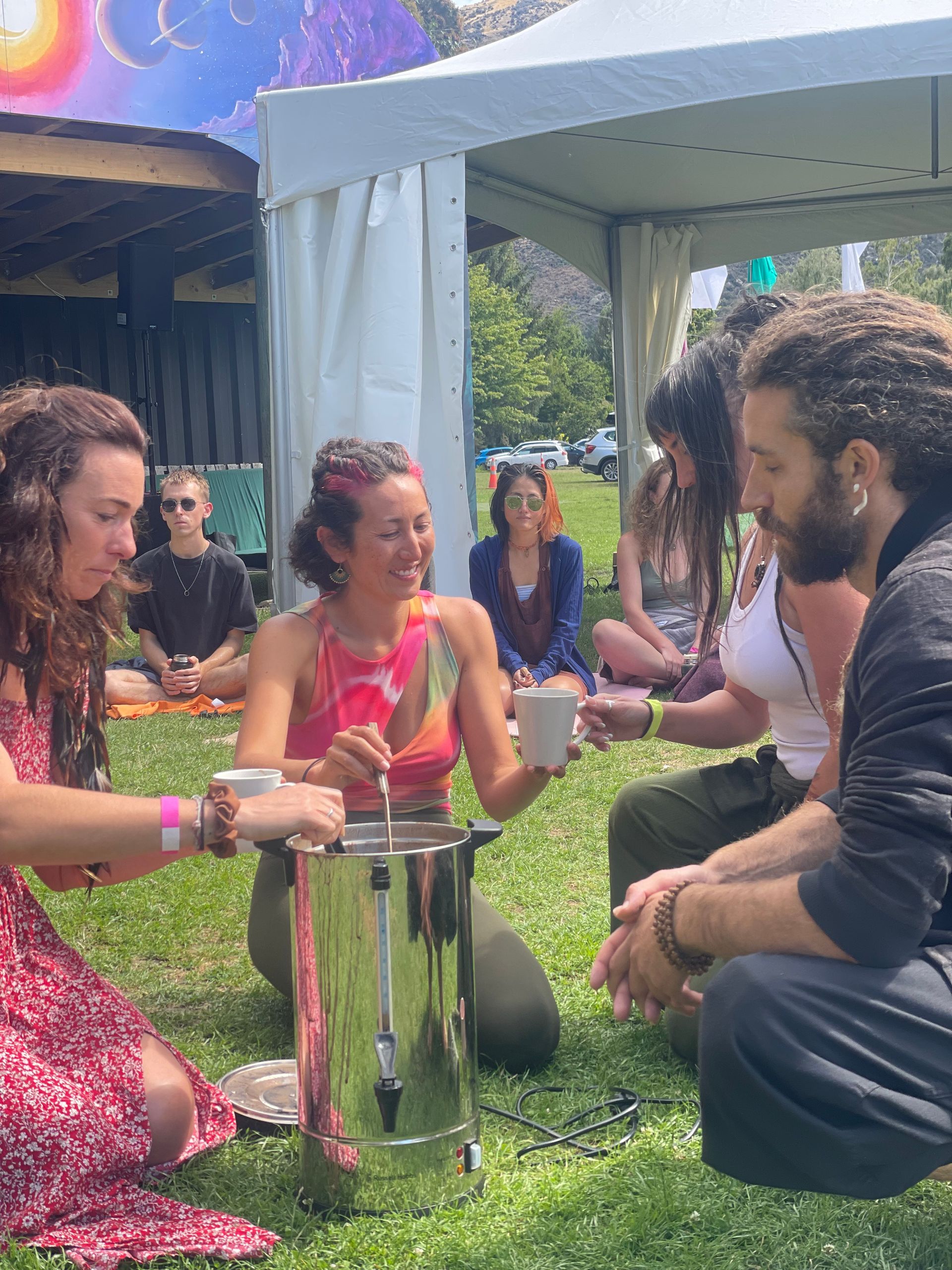 People gathering outdoors, drinking from mugs. A woman pours from a silver urn.