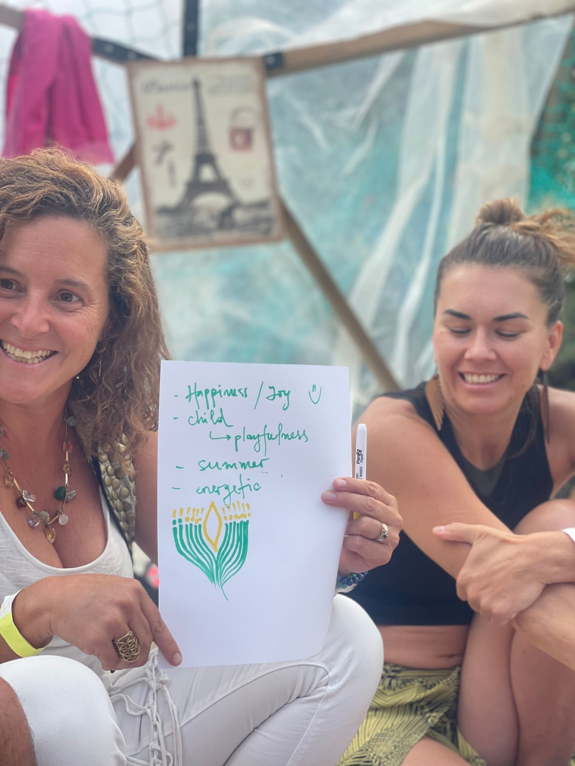 Two smiling women holding a handwritten list; one points to the text. Outdoor setting, casual attire.