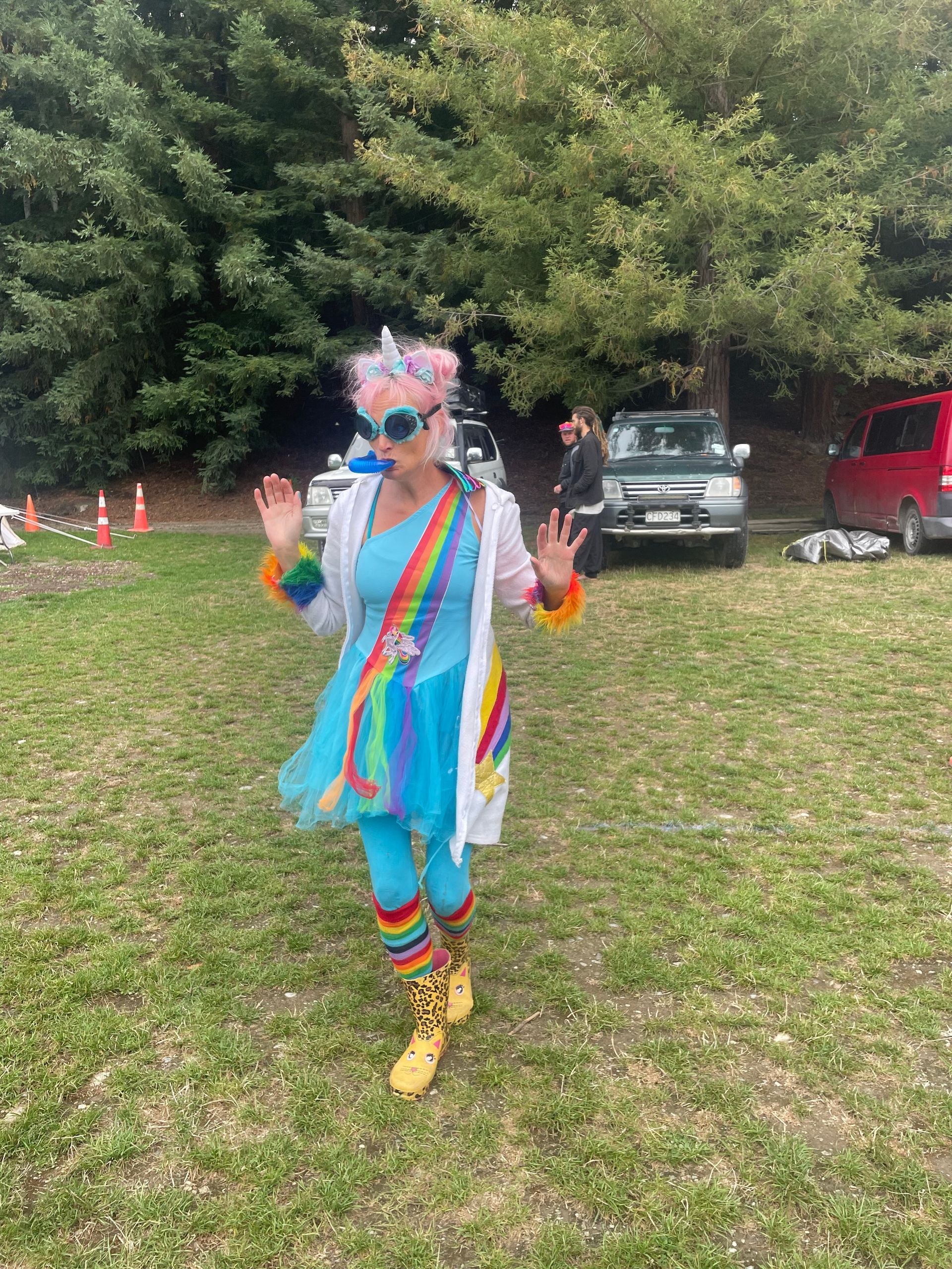Woman in a rainbow unicorn costume gesturing outdoors with cars in the background.