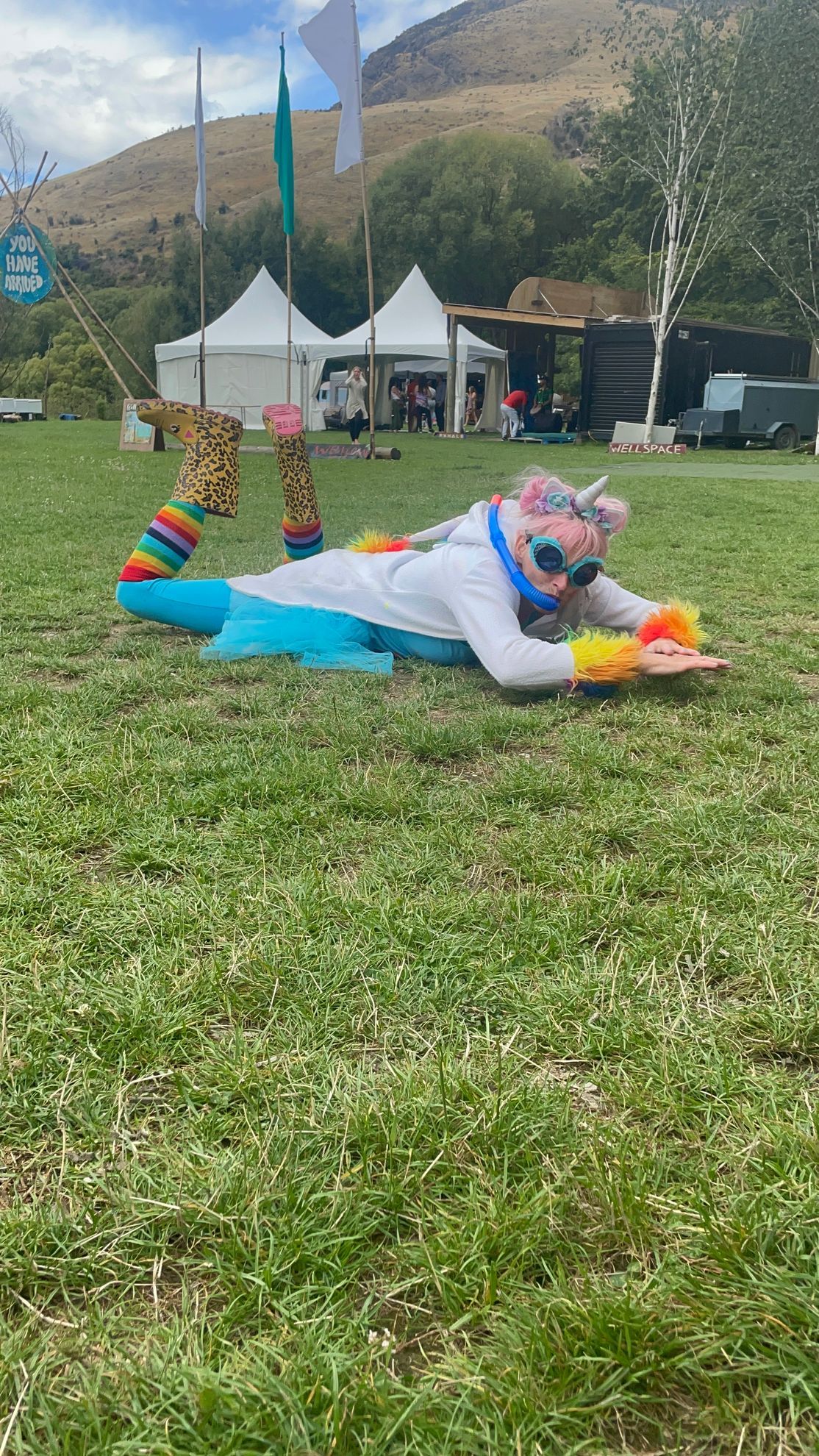 Person in costume doing a belly flop on grass at an outdoor event, rainbow colors and sunglasses, tents in the background.