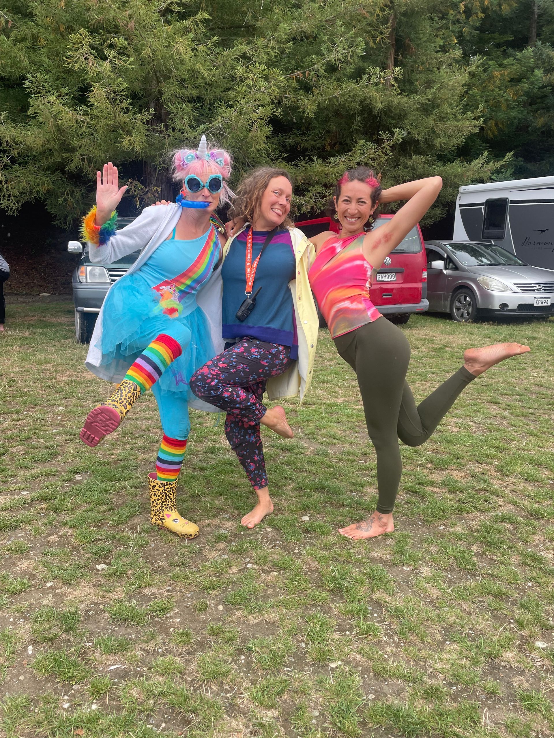 Three barefoot people pose outdoors. One in a rainbow outfit waves; the others smile with arms around each other.