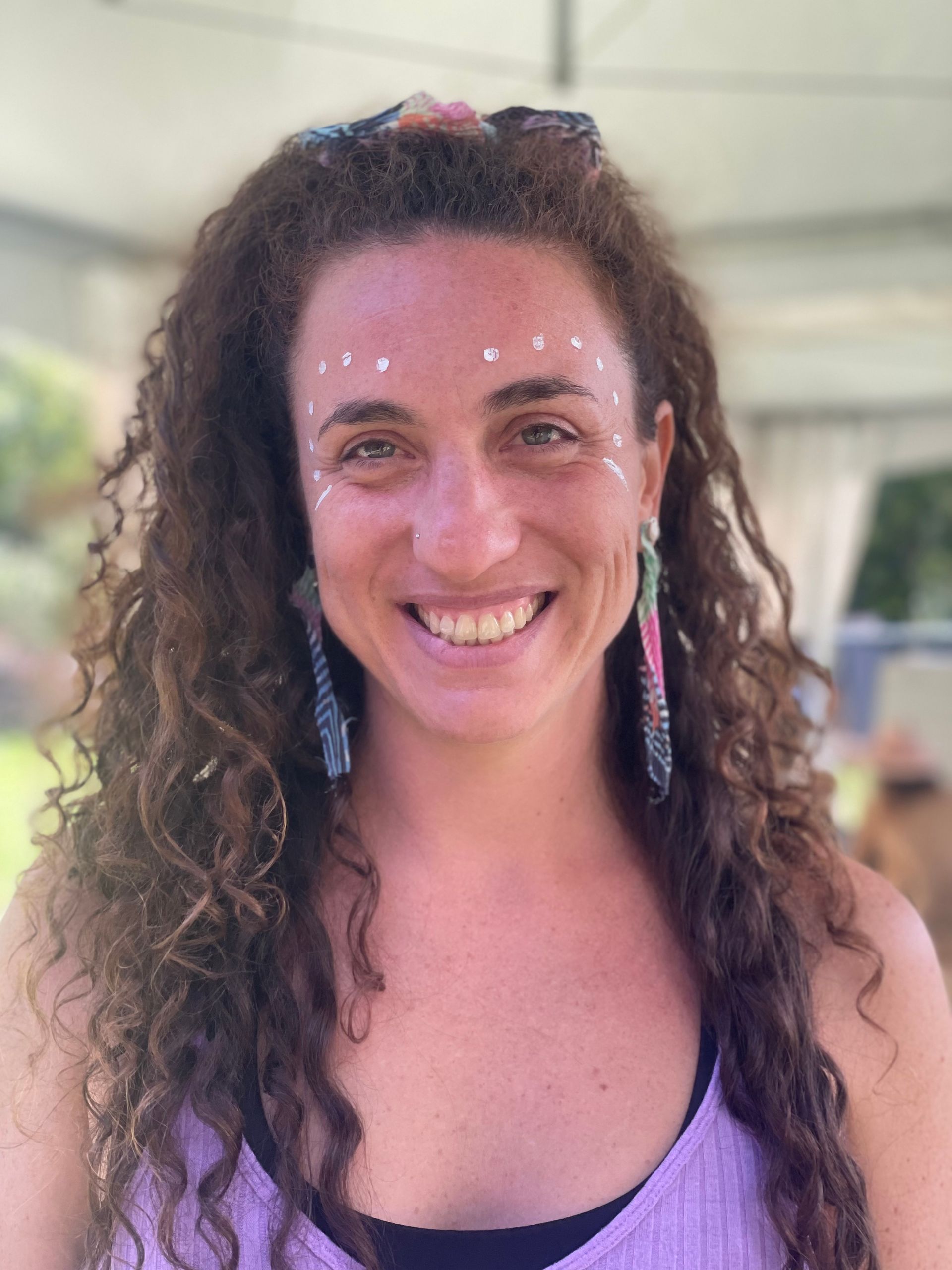 Woman with curly hair, white dots on face, smiling, wearing earrings, outdoors.