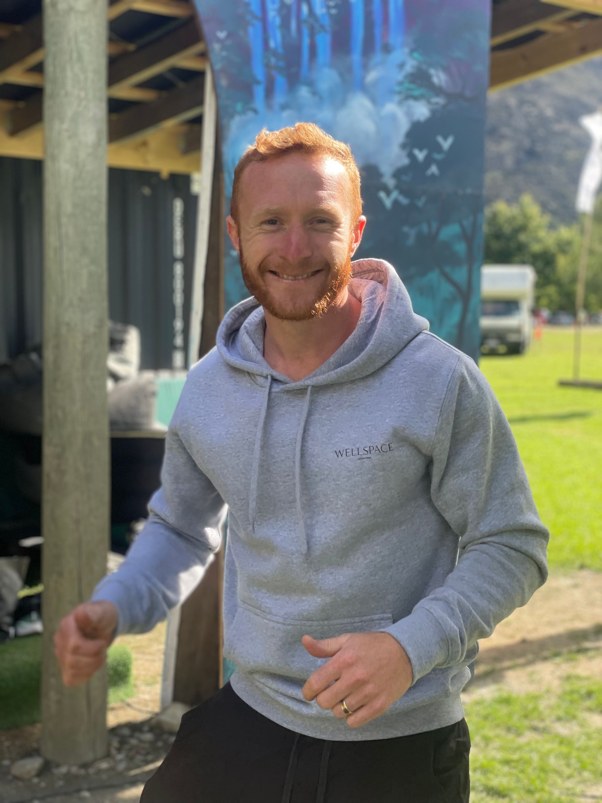 Man with red hair, wearing a gray hoodie, gives a thumbs up. Outdoors, green grass and a campervan visible.