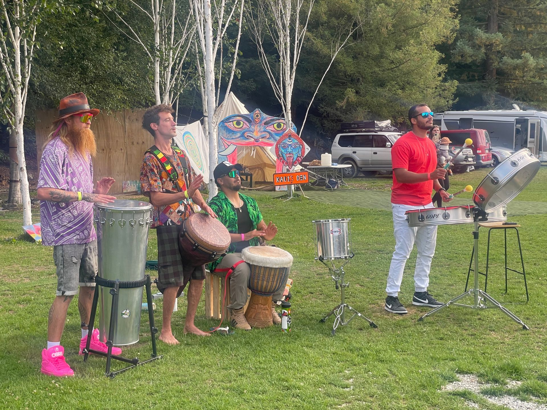 A band plays drums outdoors. The four musicians stand on grass, near trees and a tent.
